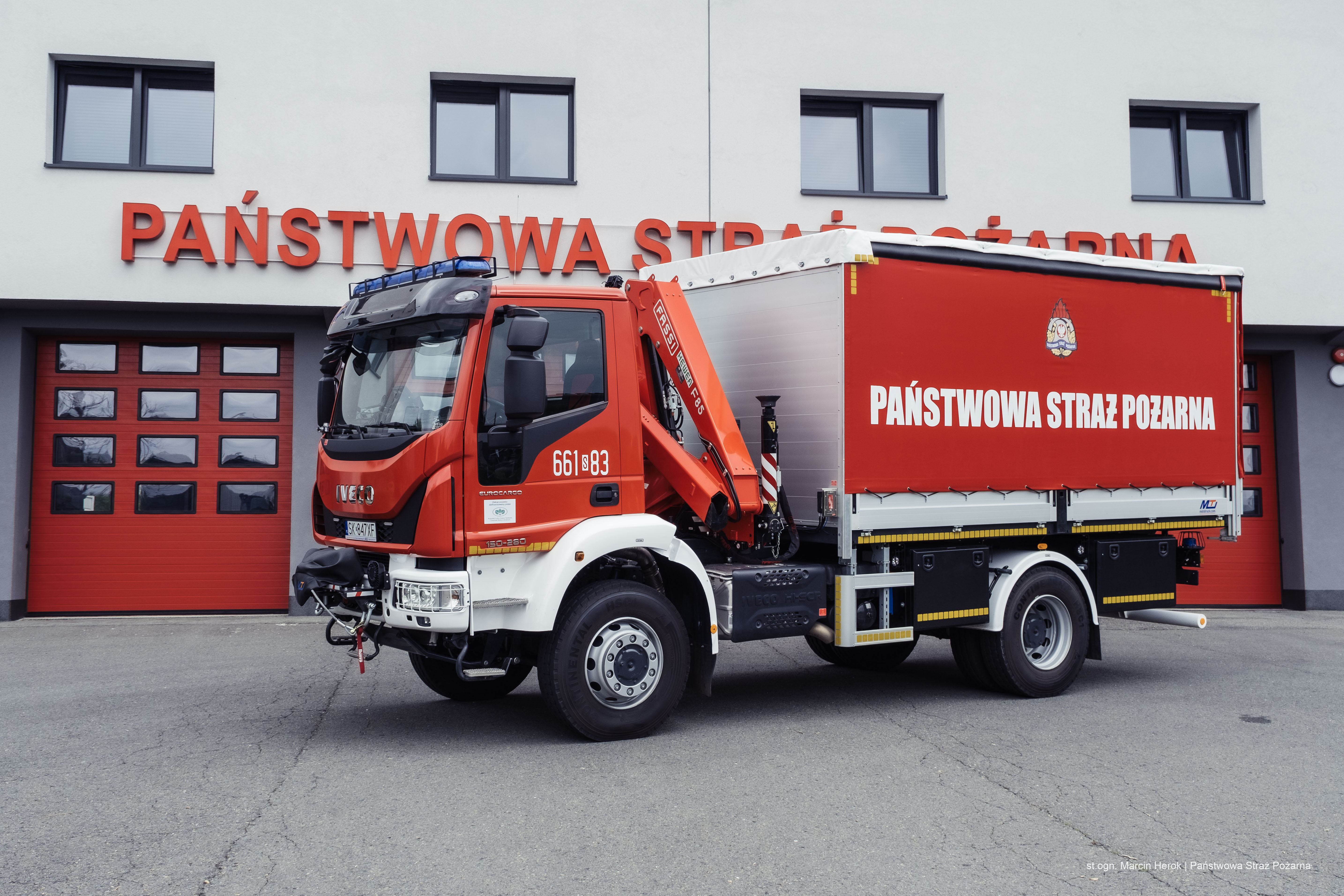 Zdjęcie przedstawia samochód kwatermistrzowski z HDS. W tle budynek Państwowej Straży Pożarnej.