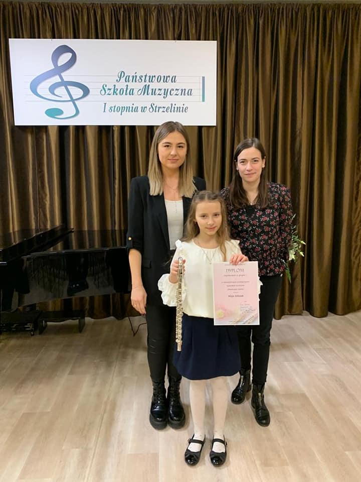 Na fotografii znajduje się uczennica Alicja Sobczak wraz z nauczycielkami: Agatą Trzepizur i Grażyną Bukartyk. Uczennica trzyma flet oraz dyplom konkursowy. W tle widoczny jest czarny fortepian oraz wiszący na ścianie baner PSM Strzelin.