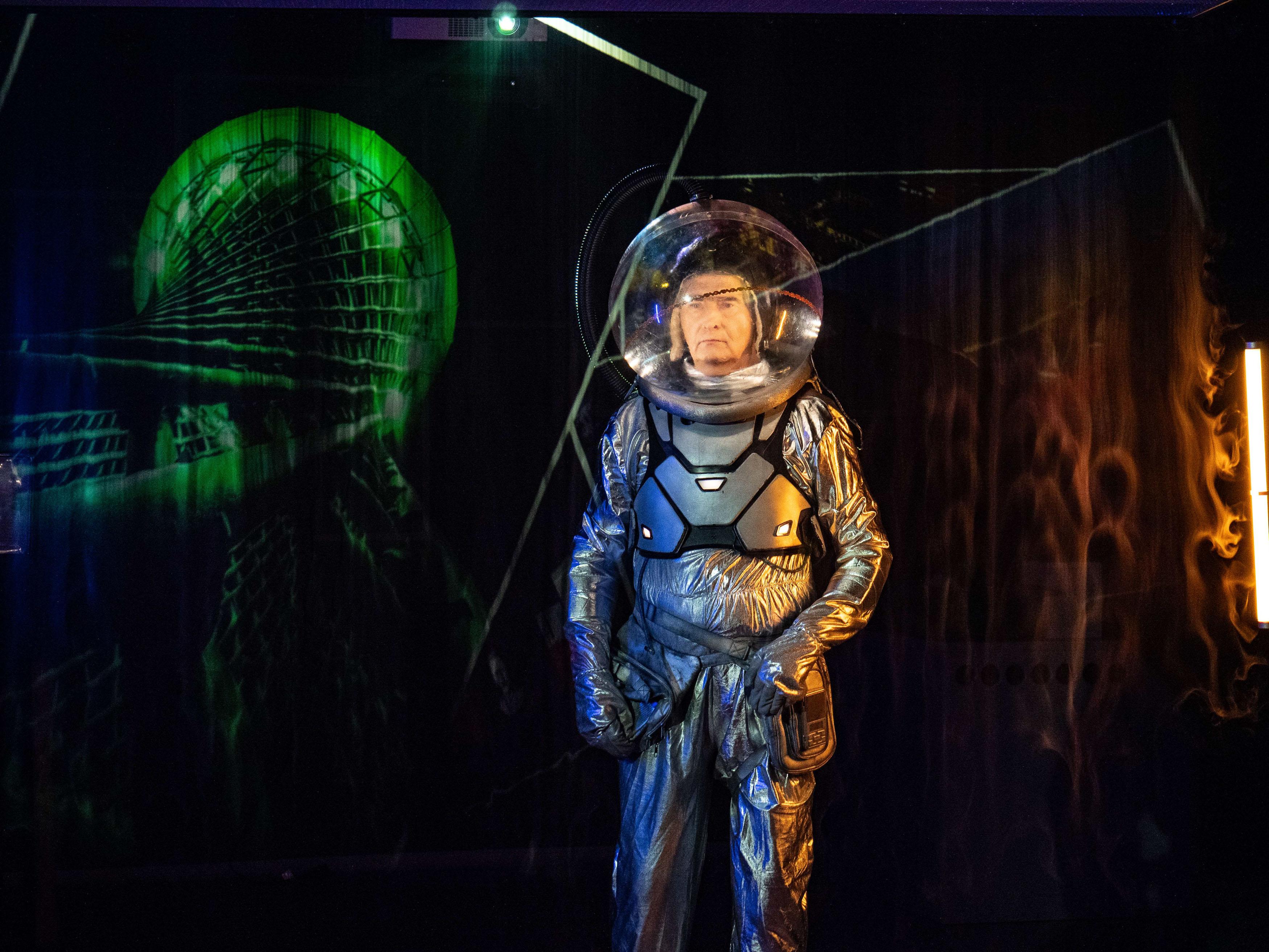 Mężczyzna w stroju astronauty stoi w ciemnym pomieszczeniu, gdzie na ścianach wyświetlają się kolorowe figury, wzory