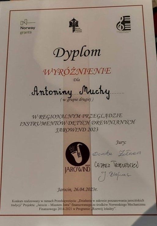 Zdjęcie ma białe tło. Nazwisko wyróżnionej uczennicy znajduje się na środku i zostało wpisane ręcznie. Poniżej widać logo przeglądu. Po prawej stronie są podpisy jury. 