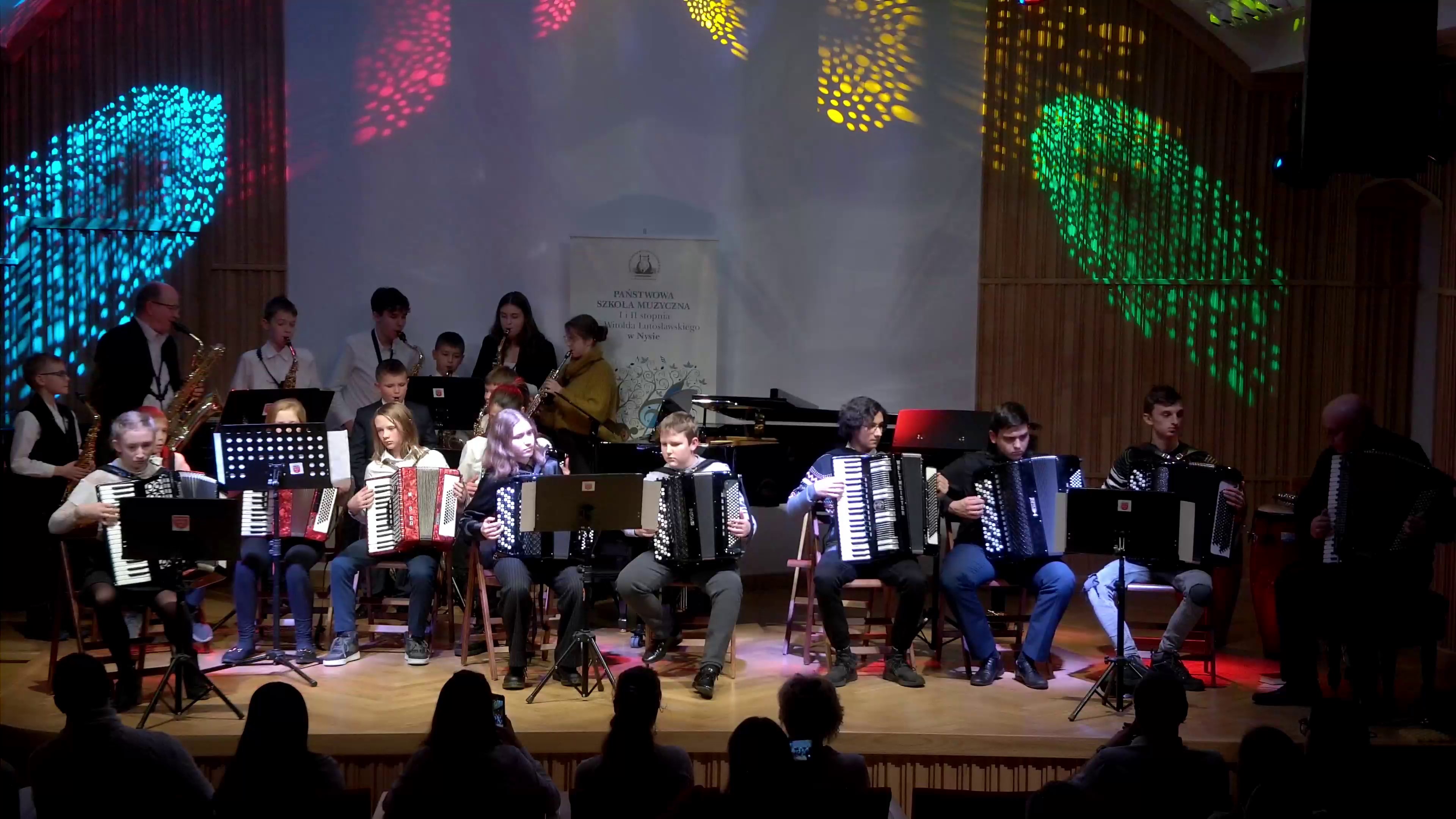 Zdjęcie. U dołu widać słuchaczy siedzących w pierwszym rzędzie widowni. Oglądają i słuchają występu zespołu akordeonistów, którzy siedzą i grają na estradzie. Za młodymi akordeonistami z lewej strony gra zespół saksofonistów. W górnej części zdjęcia widać różnokolorowe świetlne efekty.