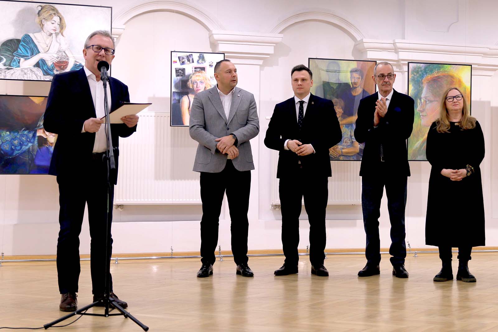 Fotografia przedstawia scenę z otwarcia wystawy. Na pierwszym planie znajduje się pięć osób stojących w szeregu na tle grafik. 