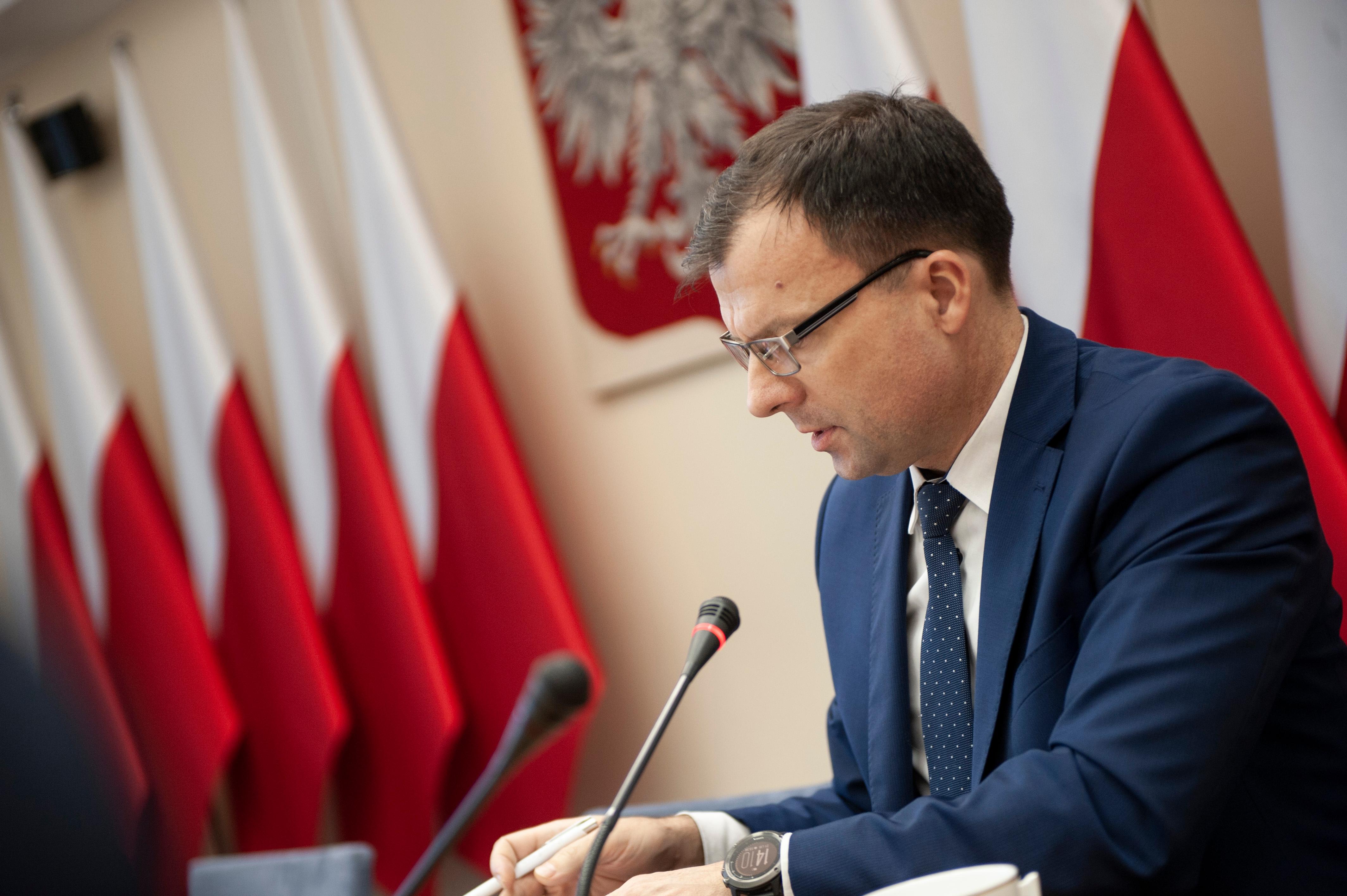 Członek komitetu mówi do mikrofonu, a za nim godło i flagi Polski.