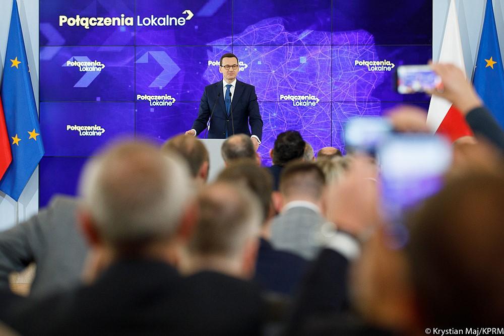 Premier Mateusz Morawiecki przemawia do siedzących naprzeciwko uczestników spotkania, a za nim ścianka wizyjna z logo Połączenia Lokalne.