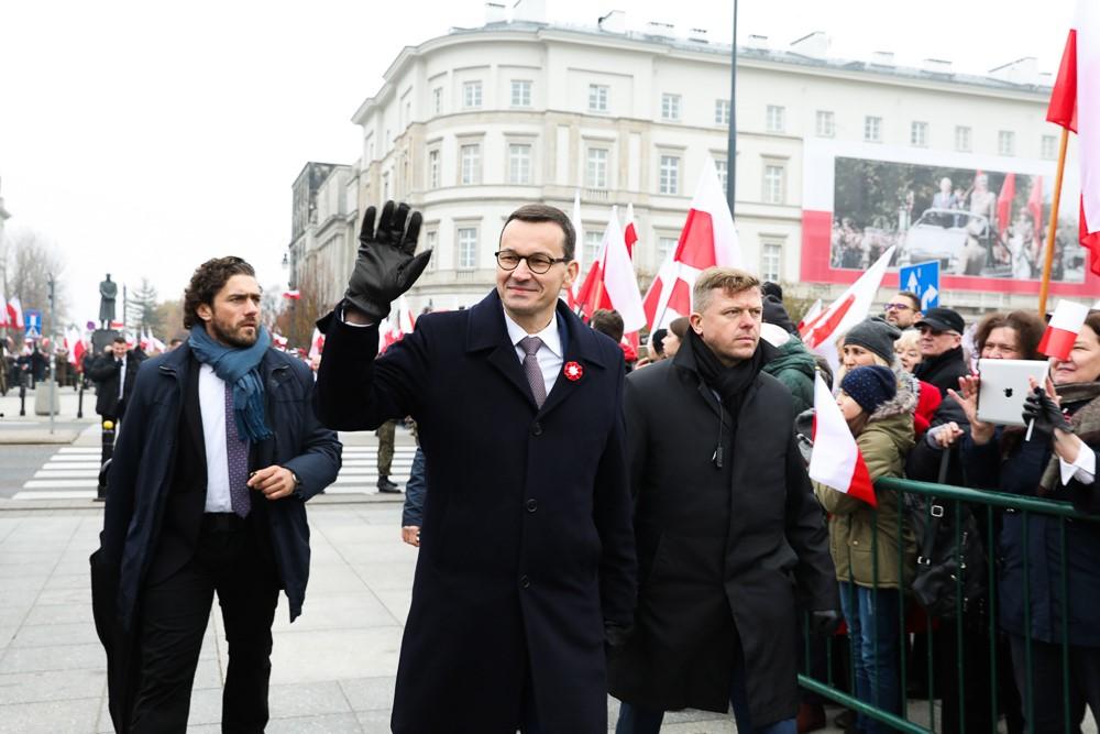 Premier Mateusz Morawiecki idzie i macha, a za nim uczestnicy marszu z flagami Polski w dłoniach.