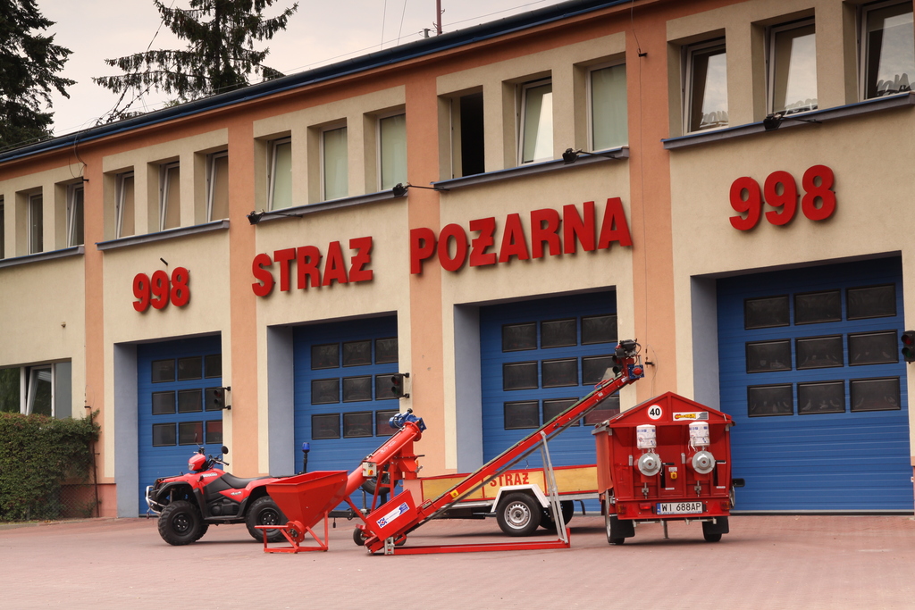 Na pierwszym tle widać zespół urządzeń składający się z podajnika ślimakowego do piasku, regulowanego taśmociągu oraz urządzenia do napełniania worków z piaskiem. Obok widoczny jest również quad z przyczepą jednoosiową do ich transportu. W tle można zobaczyć żółto-pomarańczowy budynek Jednostki Ratowniczo-Gaśniczej, czerwony napis "998 Straż Pożarna" oraz niebieskie bramy garażowe. 