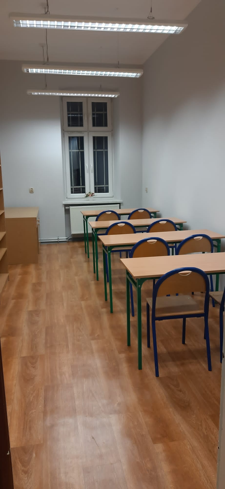 Zdjęcie przedstawia pomieszczenie, w której znajduje się czytelnia. Widoczny jest rząd podwójnych ławek wraz z krzesłami. Na zdjęciu widoczne jest także okno oraz wiszące lampy.