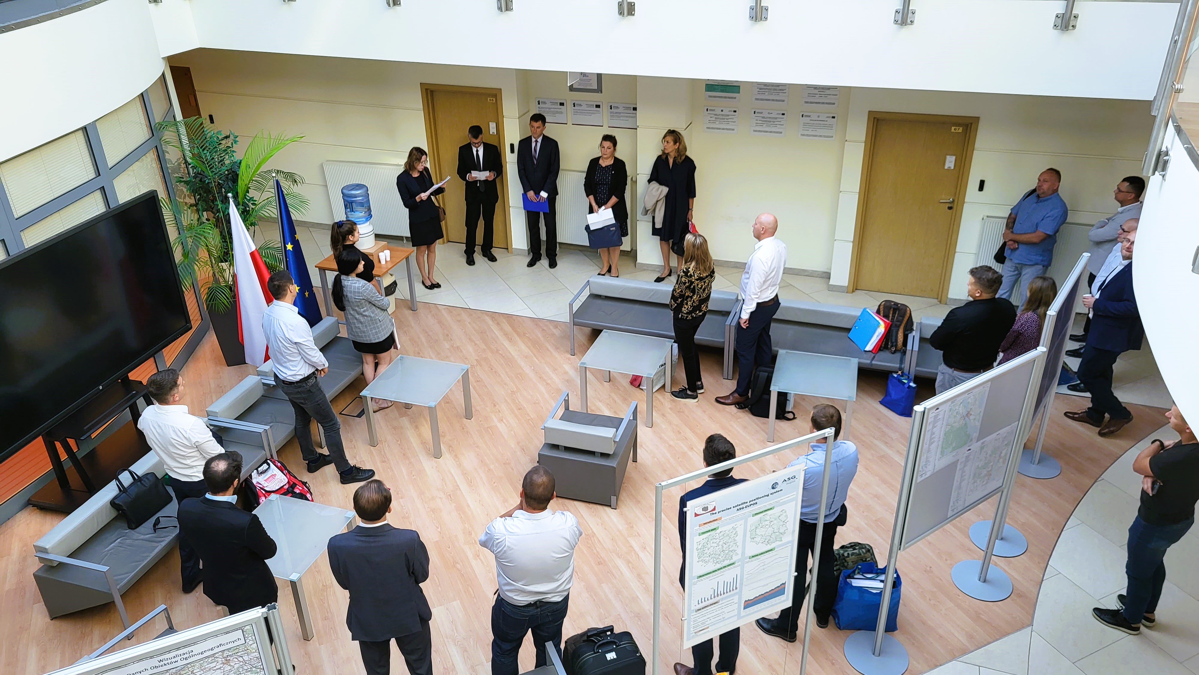 Zdjęcie przedstawia hol w siedzibie GUGiK przy ul. Olbrachta 94B z osobami przystępującymi do egzaminu słuchającymi wyjaśnień członków Komisji Egzaminacyjnej. 