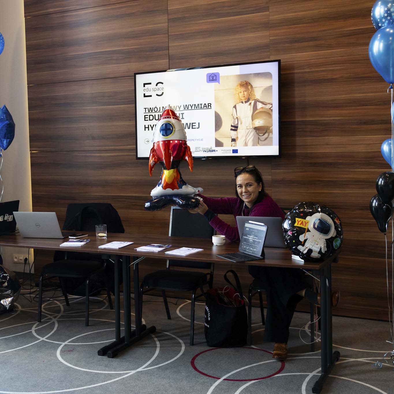 Kobieta trzyma w rękach balon w kształcenie rakiety, uśmiecha się. Obok na laptopie pracuje mężczyzna. Na około nich sa kolorowe baloniki.