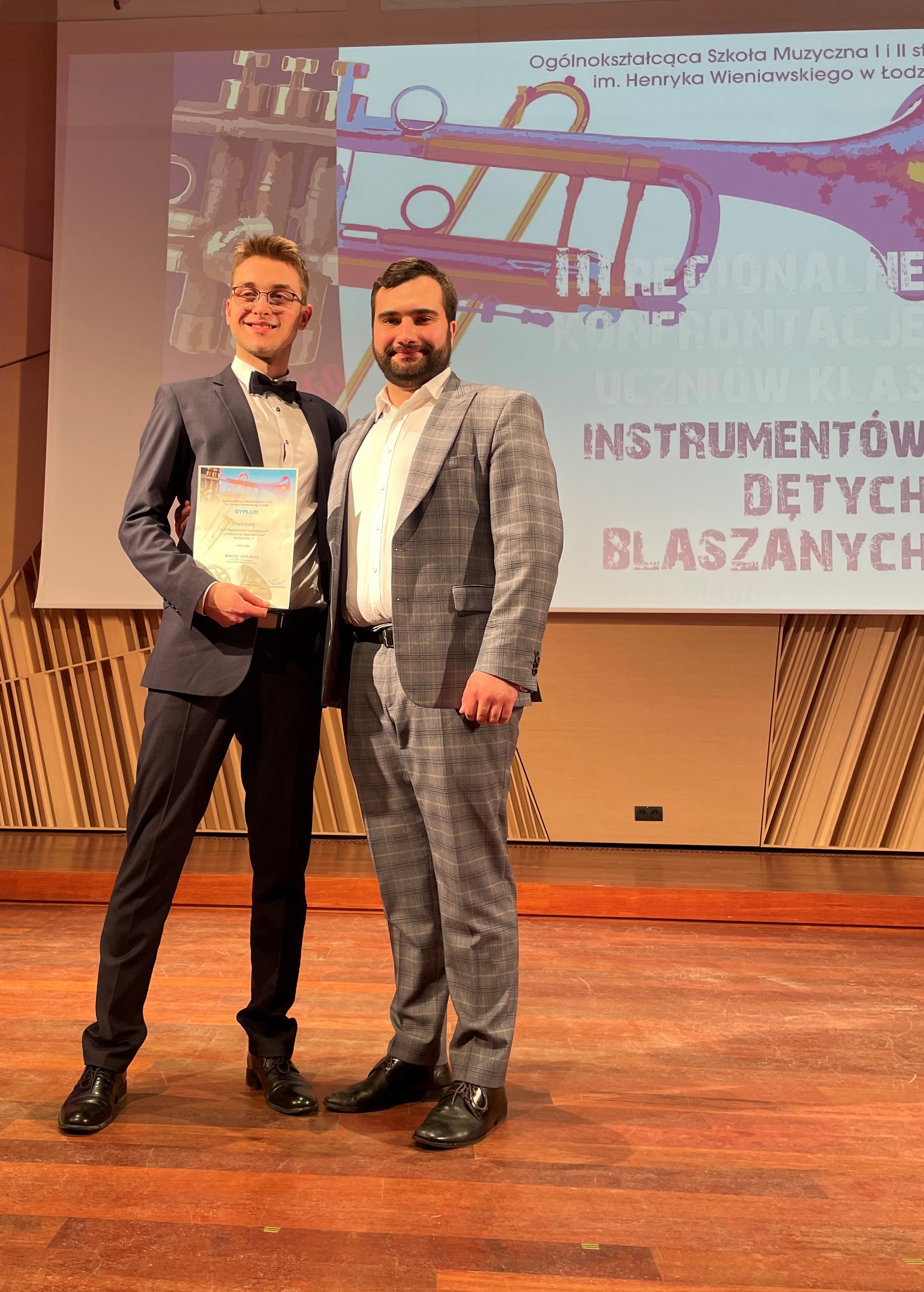 Zdjęcie przedstawia ucznia wraz z nauczycielem, którzy brali udział w konkursie "Instrumenty Dęte Blaszane" w Łodzi. Obaj panowie ubrani są w eleganckie garnitury.