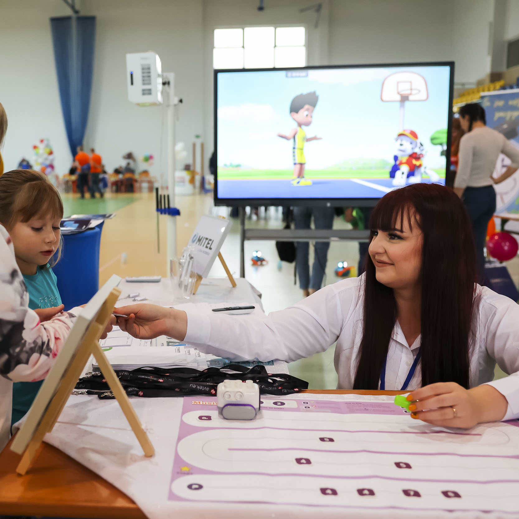 Opiekunka warsztatów podająca klocki dzieciom w trakcie zajęć, za nią ekran z wyświetlaną kreskówką