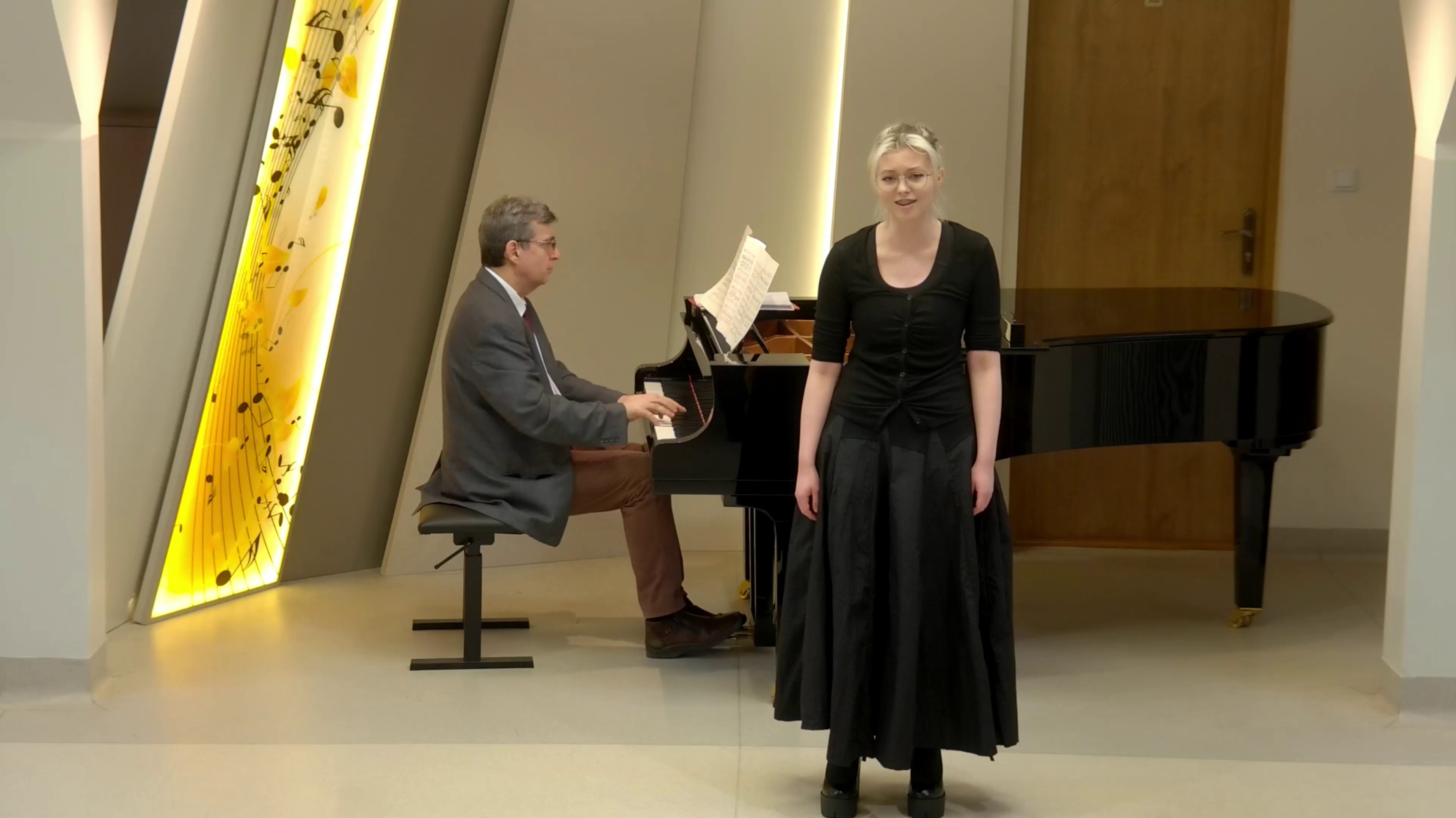 Zdjęcie. W sali kameralnej PSM I i II stopnia w Nysie występuje śpiewaczka z towarzyszeniem pianisty grającego na dużym czarnym fortepianie. W tle wyświetlony baner z nutkami i stylizowana pięciolinią.
