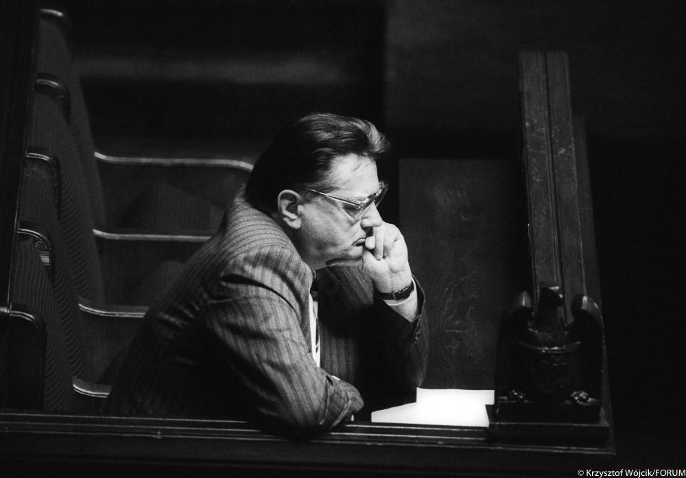 Czarno-białe zdjęcie Jana Olszewskiego, który siedzi bokiem i podpiera głowę lewą ręką.