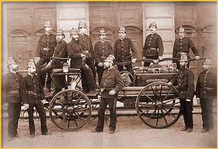 Zdjęcie historyczne w sepii przedstawiające wóz bojowy ze strażakami. 