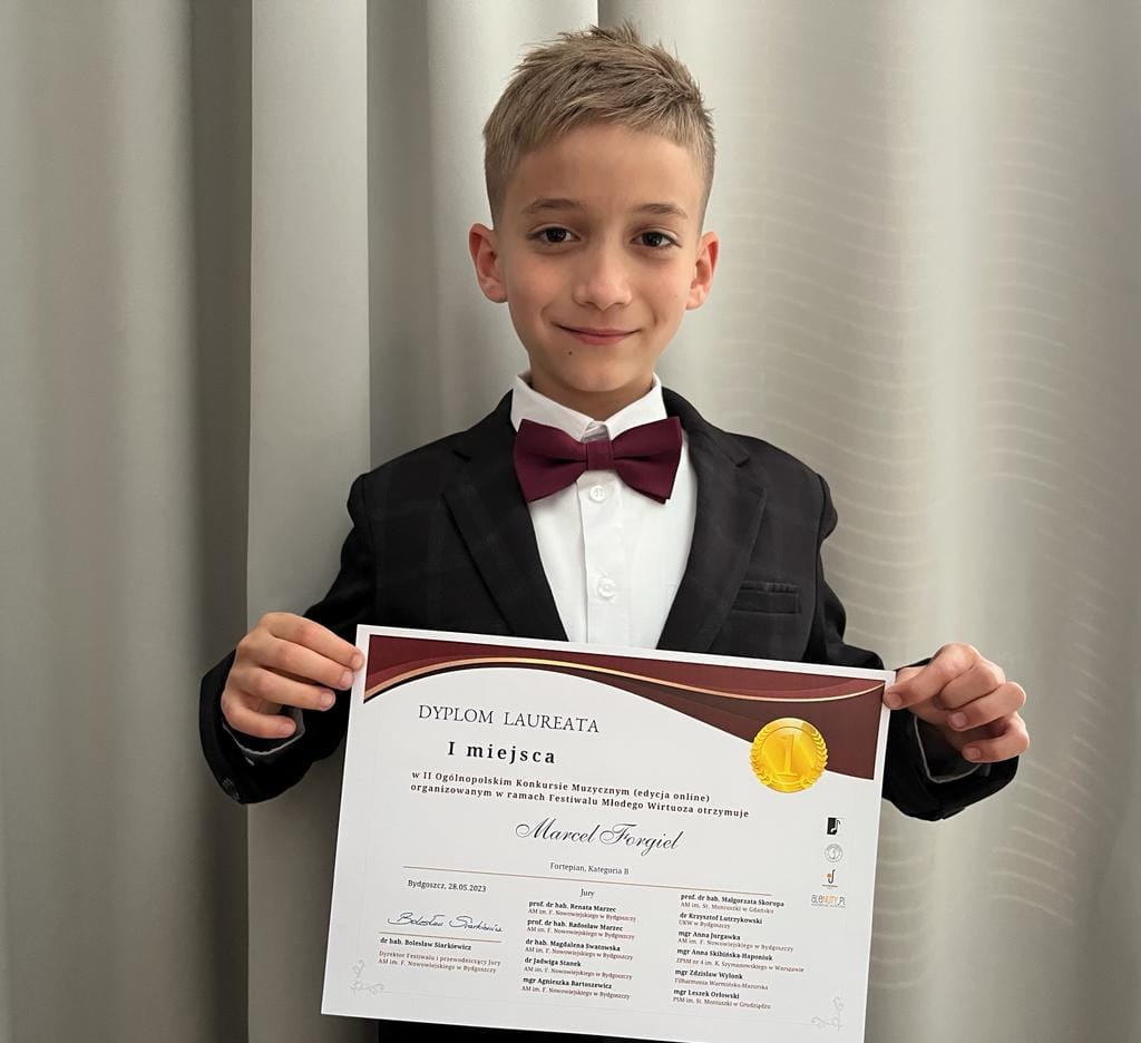 Na zdjęciu widoczny jest uśmiechnięty i elegancko ubrany chłopiec, trzymający w dłoniach dyplom konkursowy.