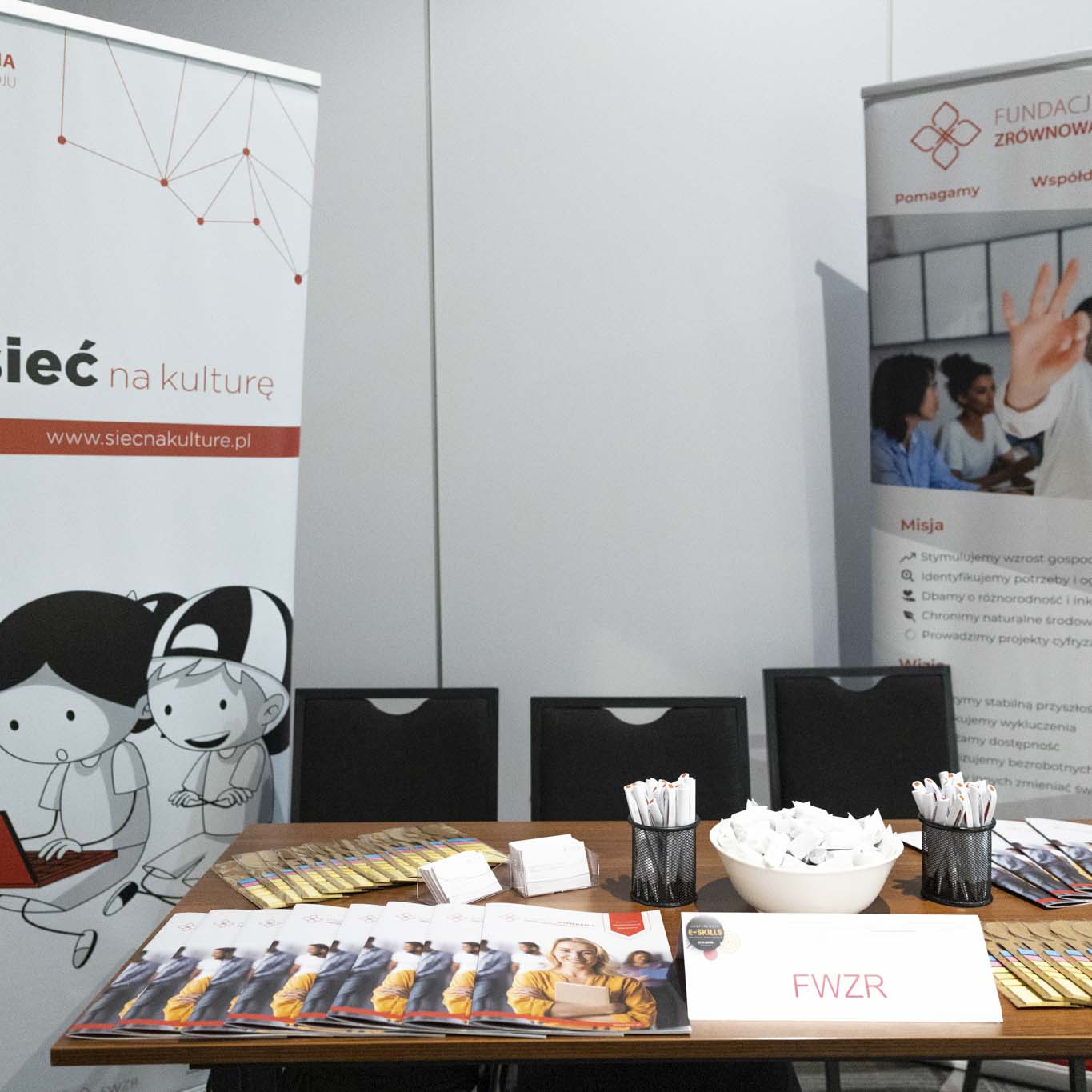 Stoisko Fundacji Wspierania Zrównoważonego Rozwoju. Biurko z krzesłami. 2 roll up'y. Na biurku leżą długopisy, foldery informacyjne, cukierki.