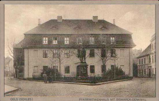 Czarno białe zdjęcie przedstawiające budynek Domu Wdów po 1910 r. od frontu w, którym aktualnie mieści się szkoła muzyczna. Przed budynkiem nieistniejący pomnik Bismarcka i dwoje stojących ludzi. Na dole fotografii napis Oles (Schles.) Pfarrwitwenhaus mit bismarck-denkmal.