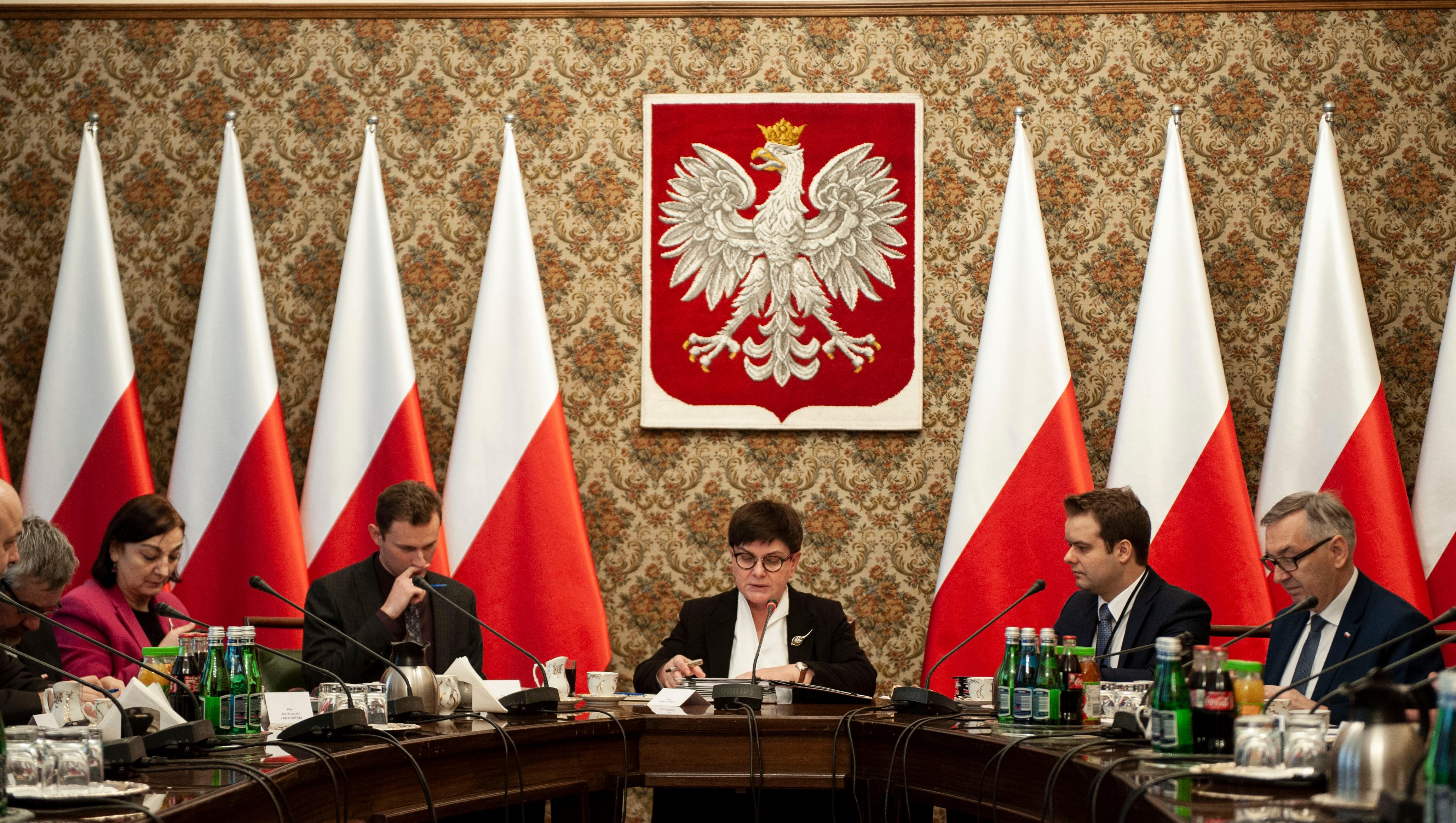Wicepremier Beata Szydło ze współpracownikami siedzi na tle godła i biało-czerwonych flag.