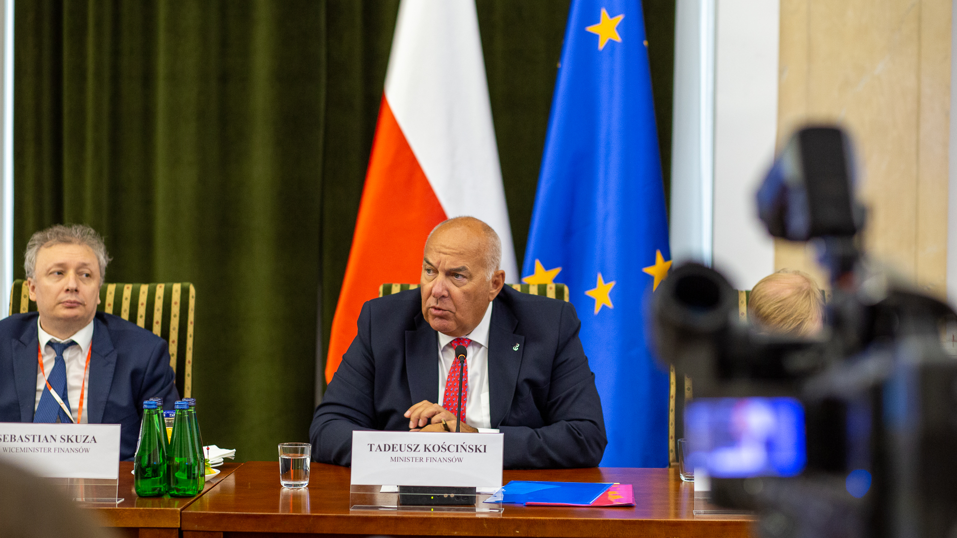 Minister finansów Tadeusz Kościński, wiceminister finansów Sebastian Skuza siedzą za stołem w tle flaga Polski i UE