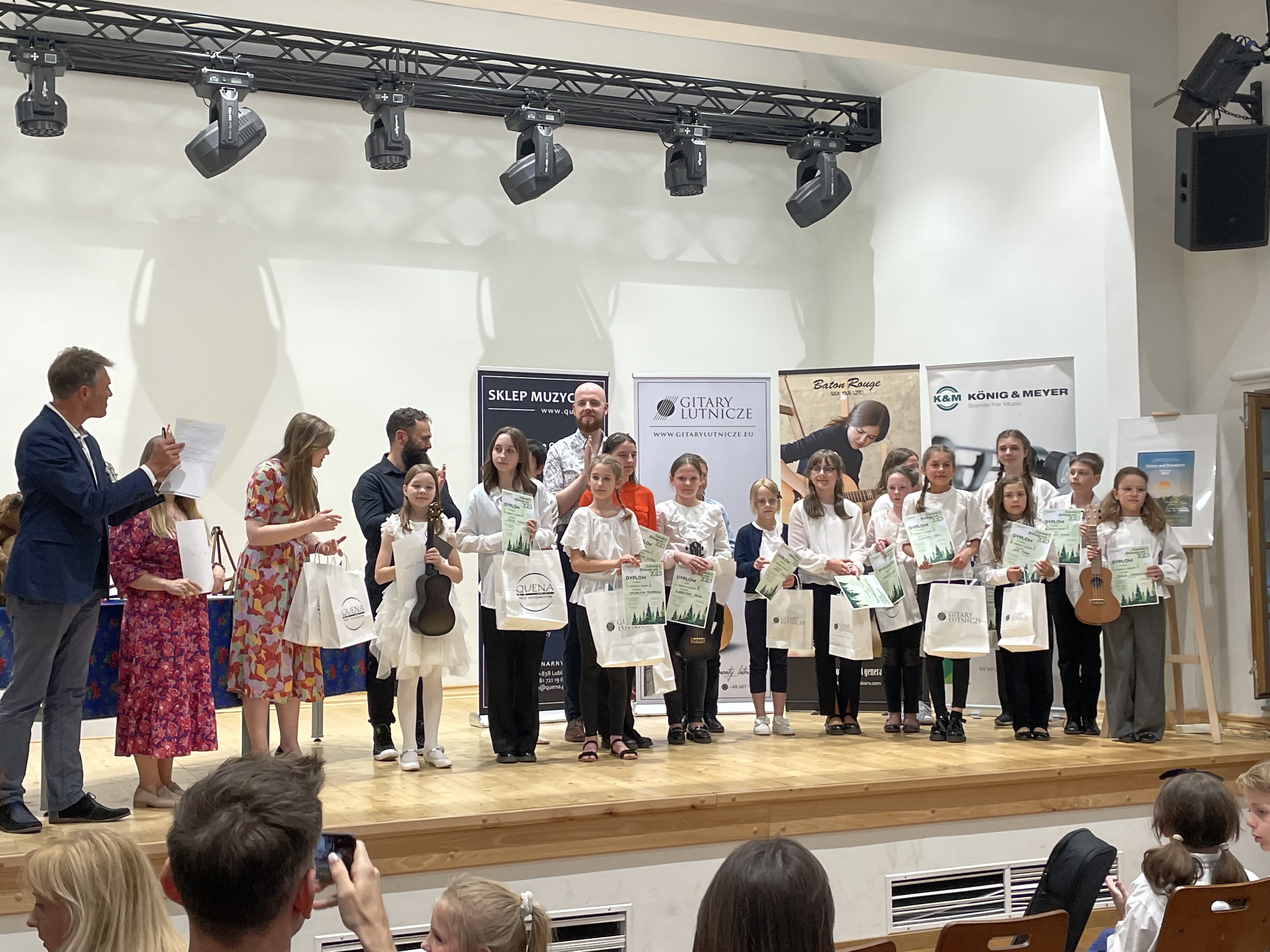 Uczennica stoi na scenie wraz z innymi laureatami konkursu, trzyma w dłoni dyplom i torbę z nagrodami. Inne dzieci również trzymają dyplomy i nagrody w torbach. Po lewej stronie stoją jurorzy konkursu oraz organizatorzy i sponsor. 