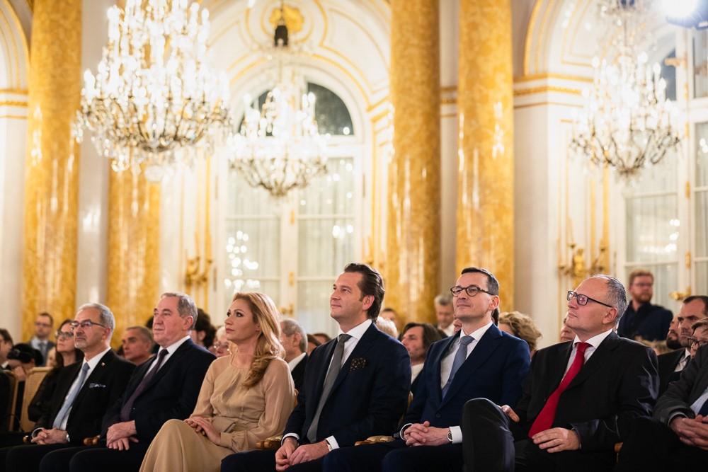 Premier Mateusz Morawiecki siedzi na widowni pośród ludzi.