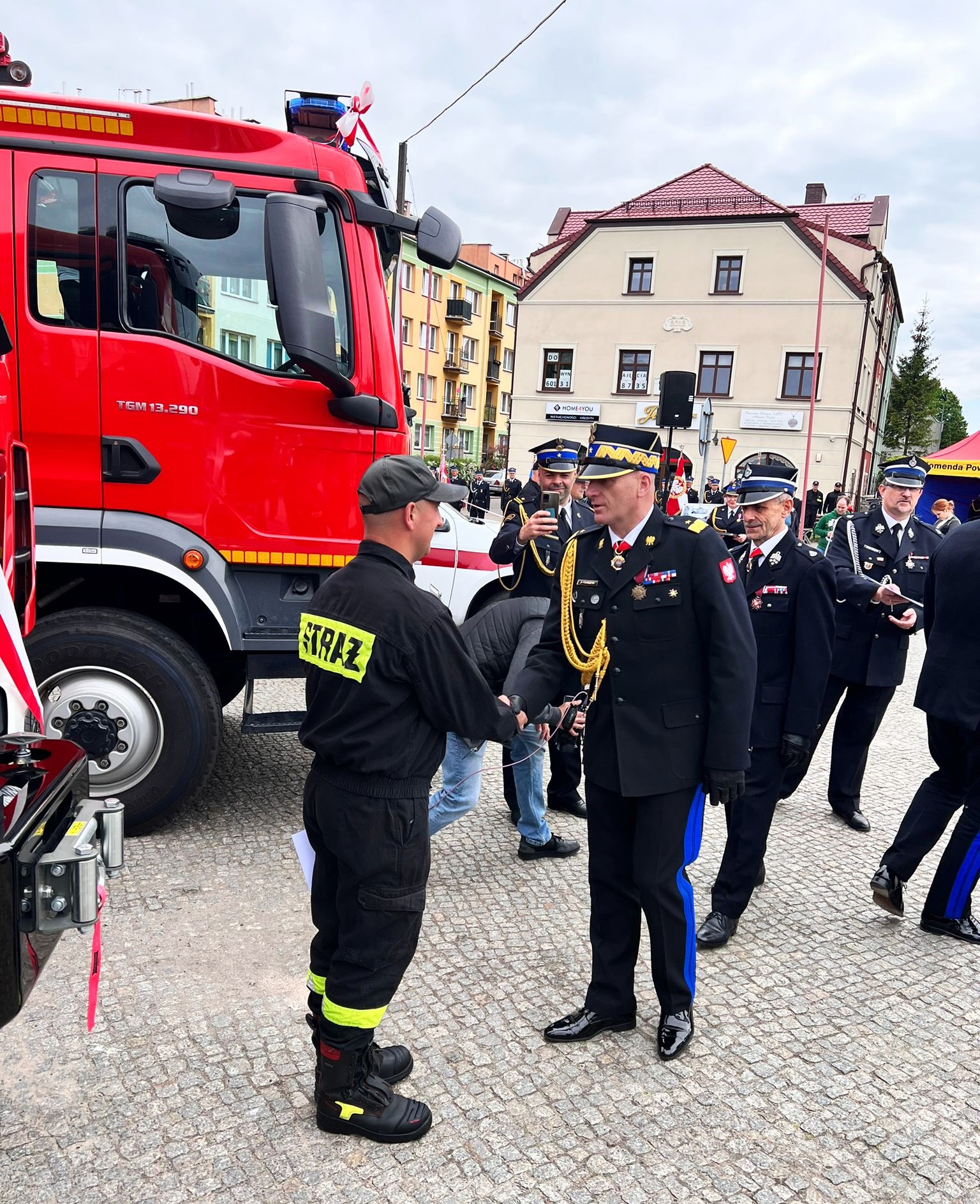 Komendant wojewódzki podaje rękę w geście gratulacji strażakowi - kierowcy, który stoi obok przekazanego samochodu ratowniczo - gaśniczego