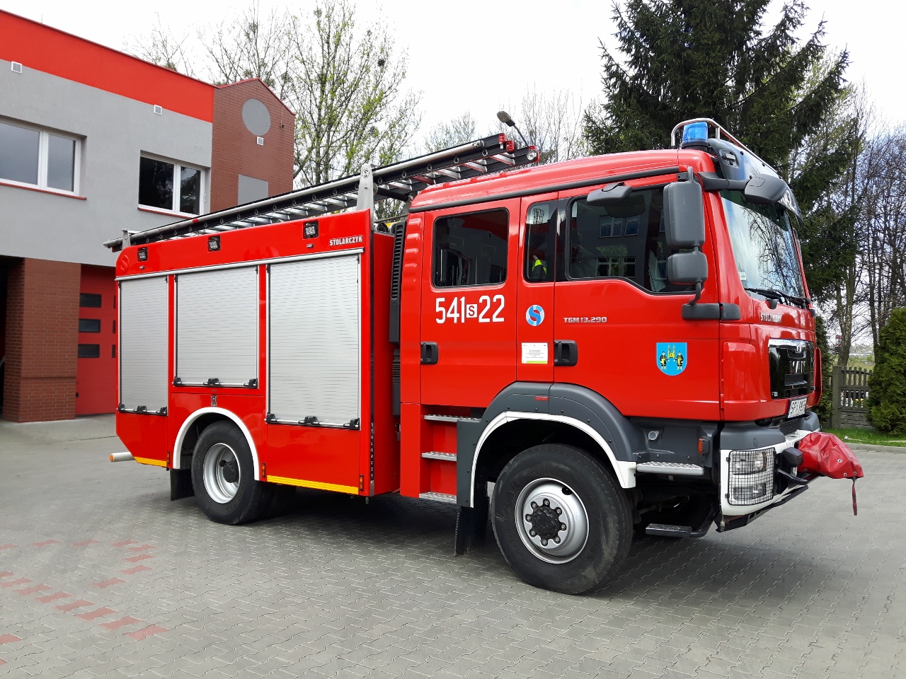 Zdjęcie przedstawia średni samochód gaśniczy marki Man w kolorze czerwonym na tle budynku komendy. w tle drzewo iglaste