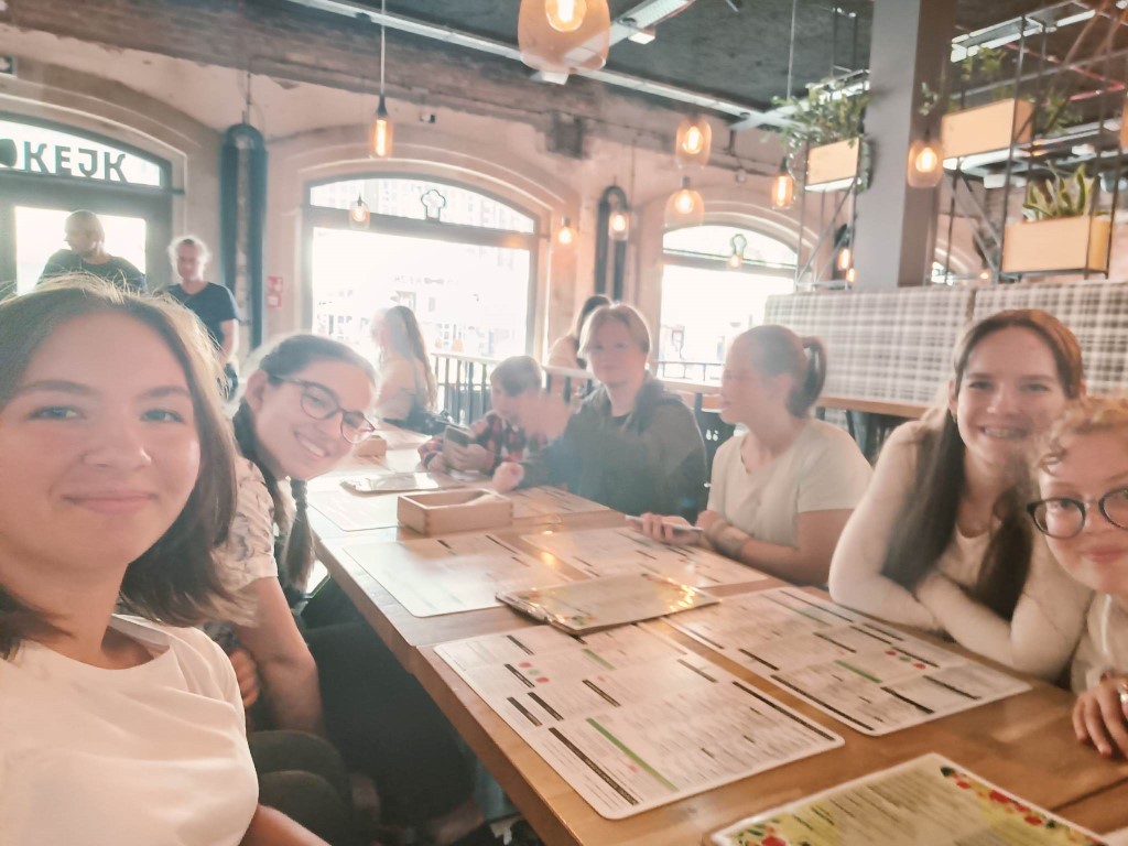 Zdjęcie kolorowe. Grupa młodzieży przy stoliku w restauracji. Na stole karty menu. W tle okno z widokiem na ulicę.