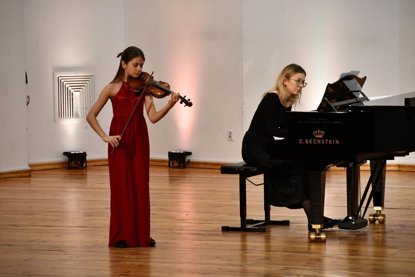 Na zdjęciu znajduje się uczennica Agata Kostanowicz grająca na skrzypcach. Ma na sobie długą, czerwoną sukienkę. Obok uczennicy znajduje się nauczycielka Pani Monika Duda, grająca na czarnym fortepianie.