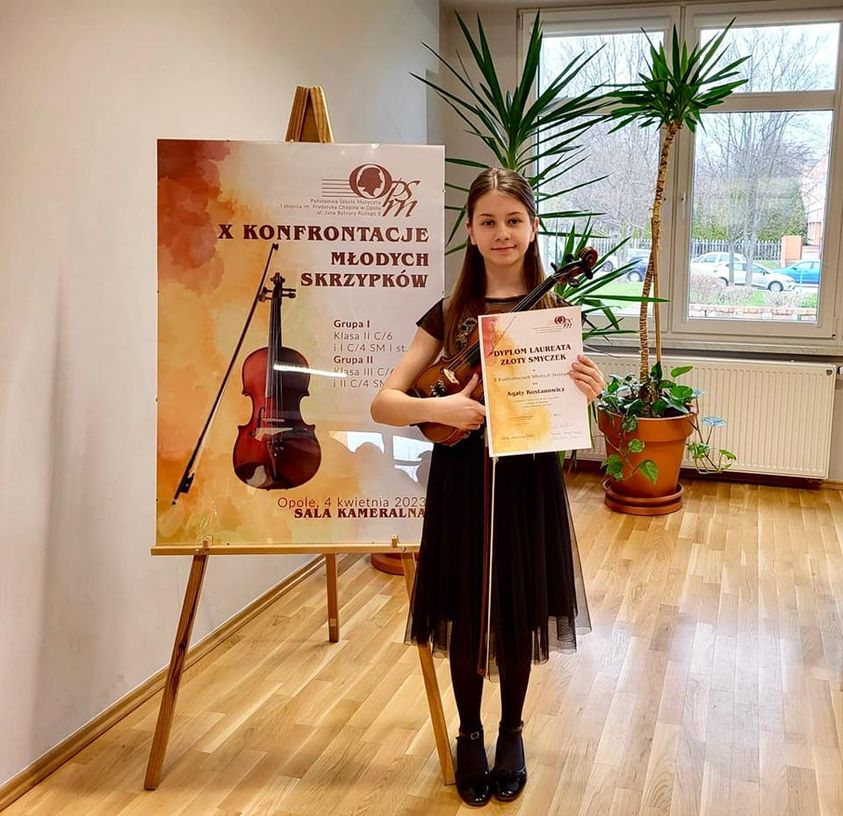 Na zdjęciu widoczna uśmiechnięta uczennica trzymająca w rękach dyplom konkursowy oraz skrzypce. Stoi na tle baneru konkursowego.