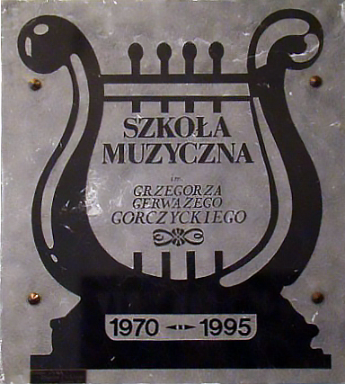 Tablica pamiątkowa przedstawiająca zarys liry z wpisanym w nią tekstem "Szkoła Muzyczna im. Grzegorza Gerwazego Gorczyckiego" oraz lata 1970-1995.