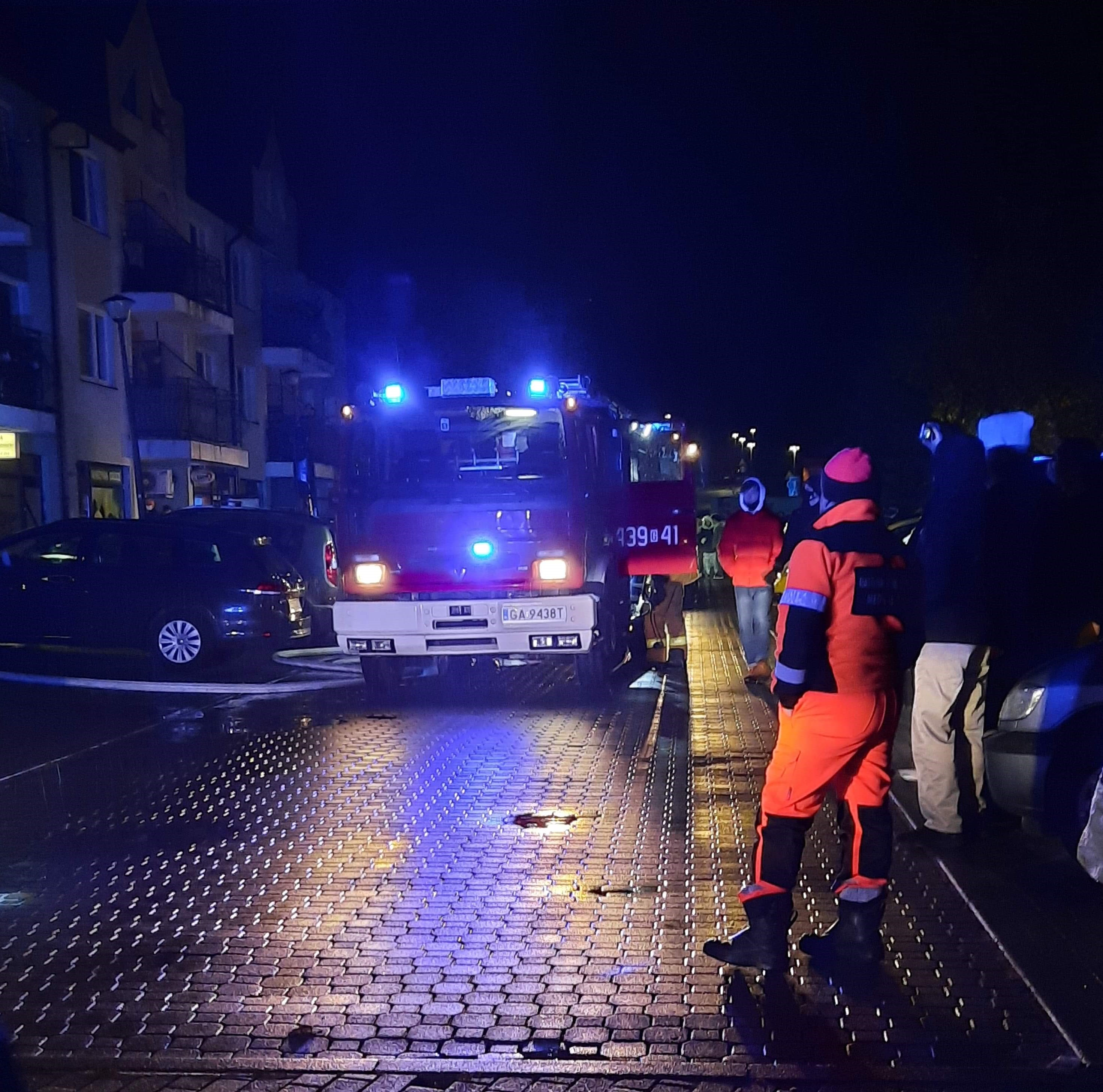 Jest ciemno. Wieczór. Czerwony wóz strażacki z włączonymi, niebieskimi światłami błyskowymi, stoi na drodze wyłożonej kostką brukową.