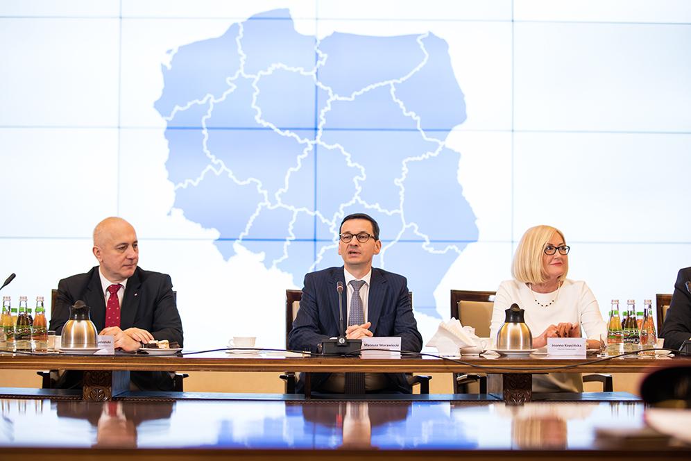 Premier Mateusz Morawiecki siedzi przy stole z uczestnikami spotkania.