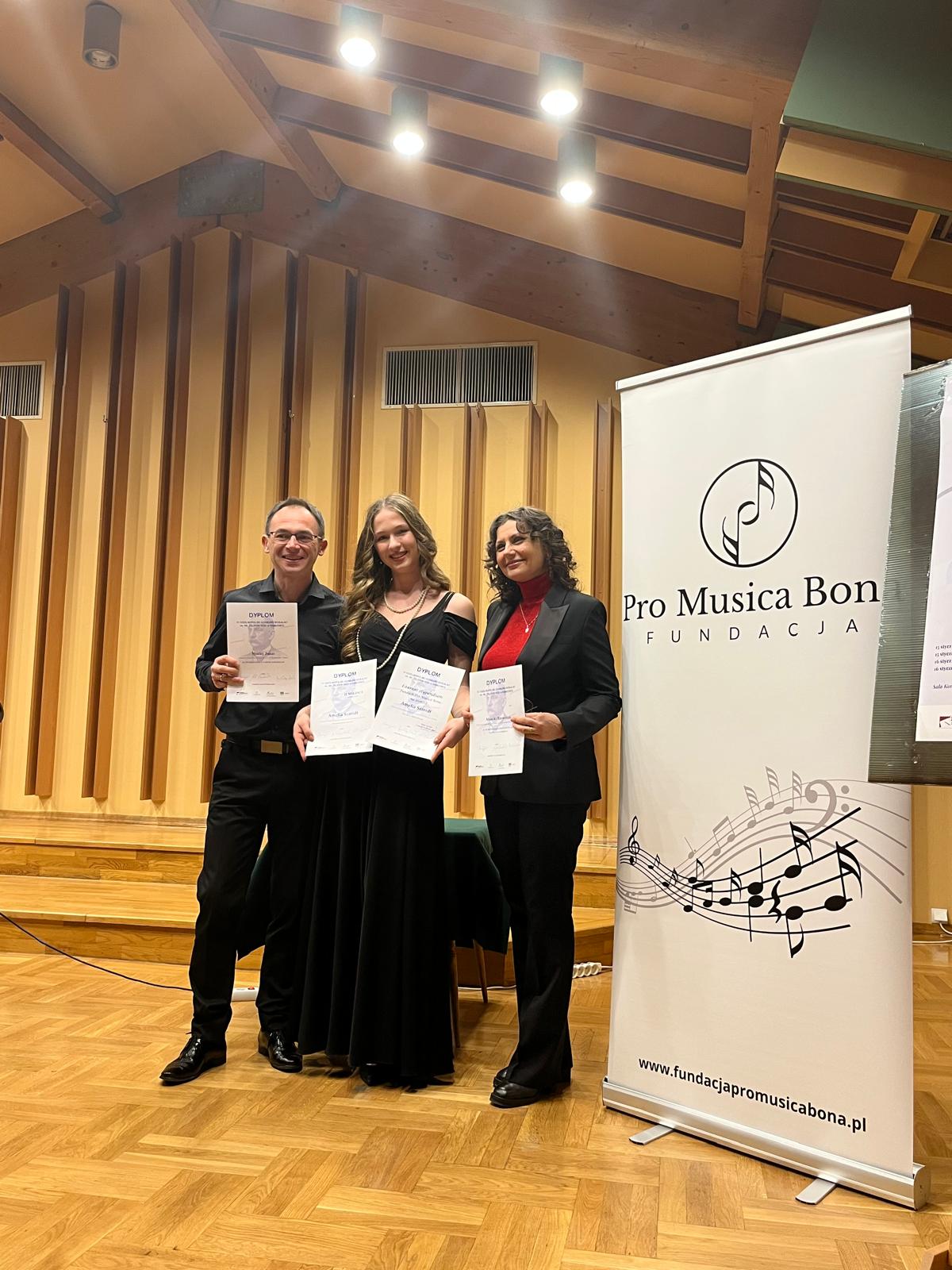 Na zdjęciu - Maciej Janas, Amelia_Szmidt, Monika_Brewczak. W tle scena konkursu i baner fundacji.