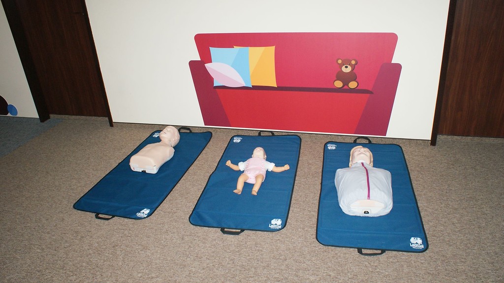 Na zdjęciu znajdują się trzy fantomy do ćwiczeń: dorosły, niemowlak i dziecko. Leżą one na podłodze na niebieskich matach. Na ścianie jest ilustracja czerwonej sofy z trzema poduszkami i pluszowym misiem.
