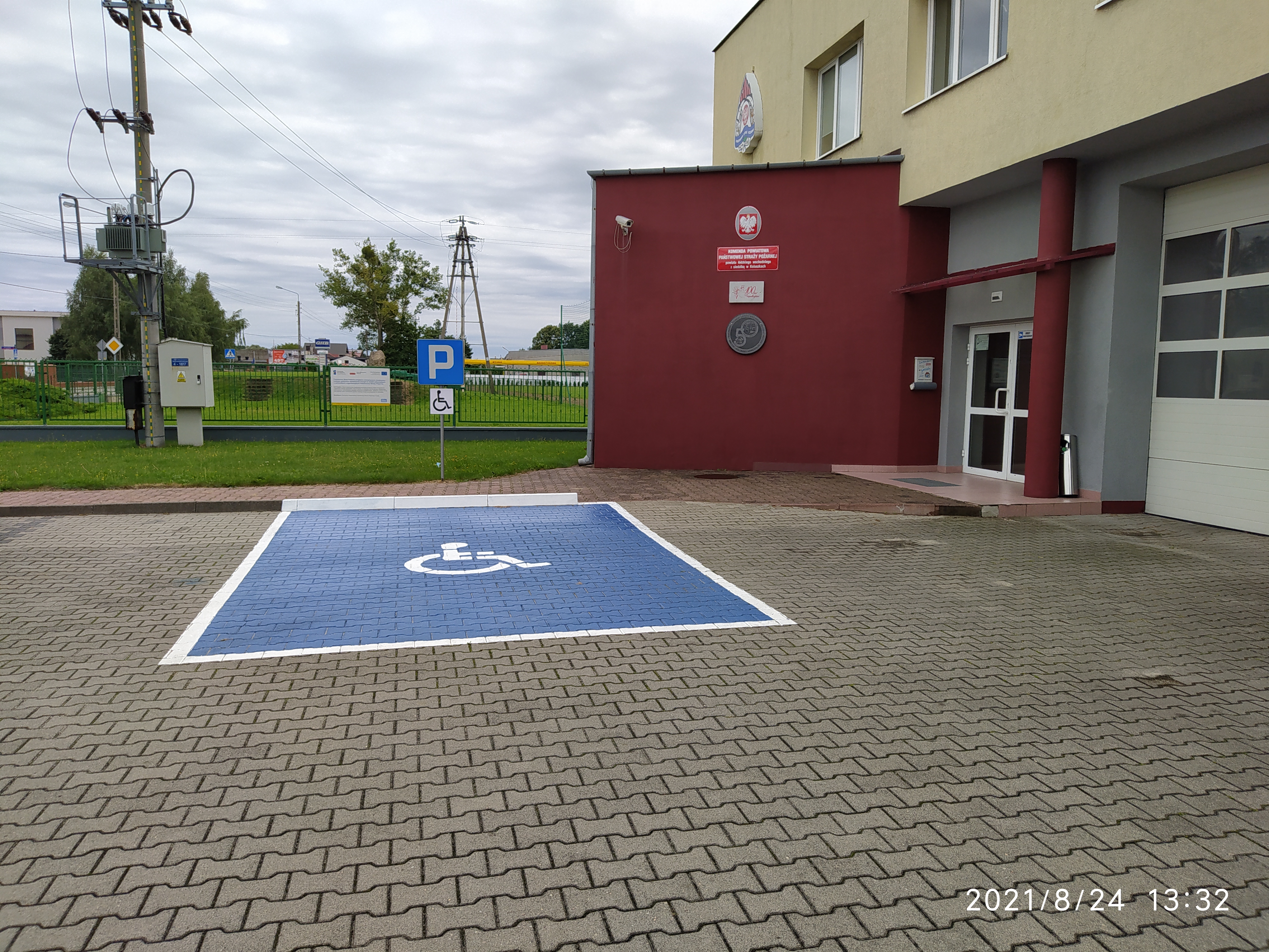 Zdjęcie przedstawia wejście do budynku Komendy Powiatowej PSP w Koluszkach, przed drzwiami widoczne miejsce parkingowe dla osób niepełnosprawnych. 