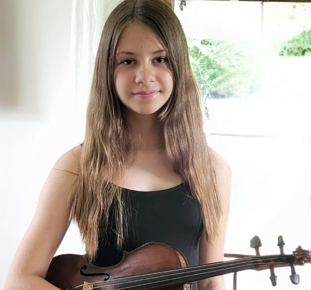Na zdjęciu znajduje się uśmiechnięta dziewczyna w rozpuszczonych włosach i czarnej bluzce. W rękach trzyma skrzypce. 