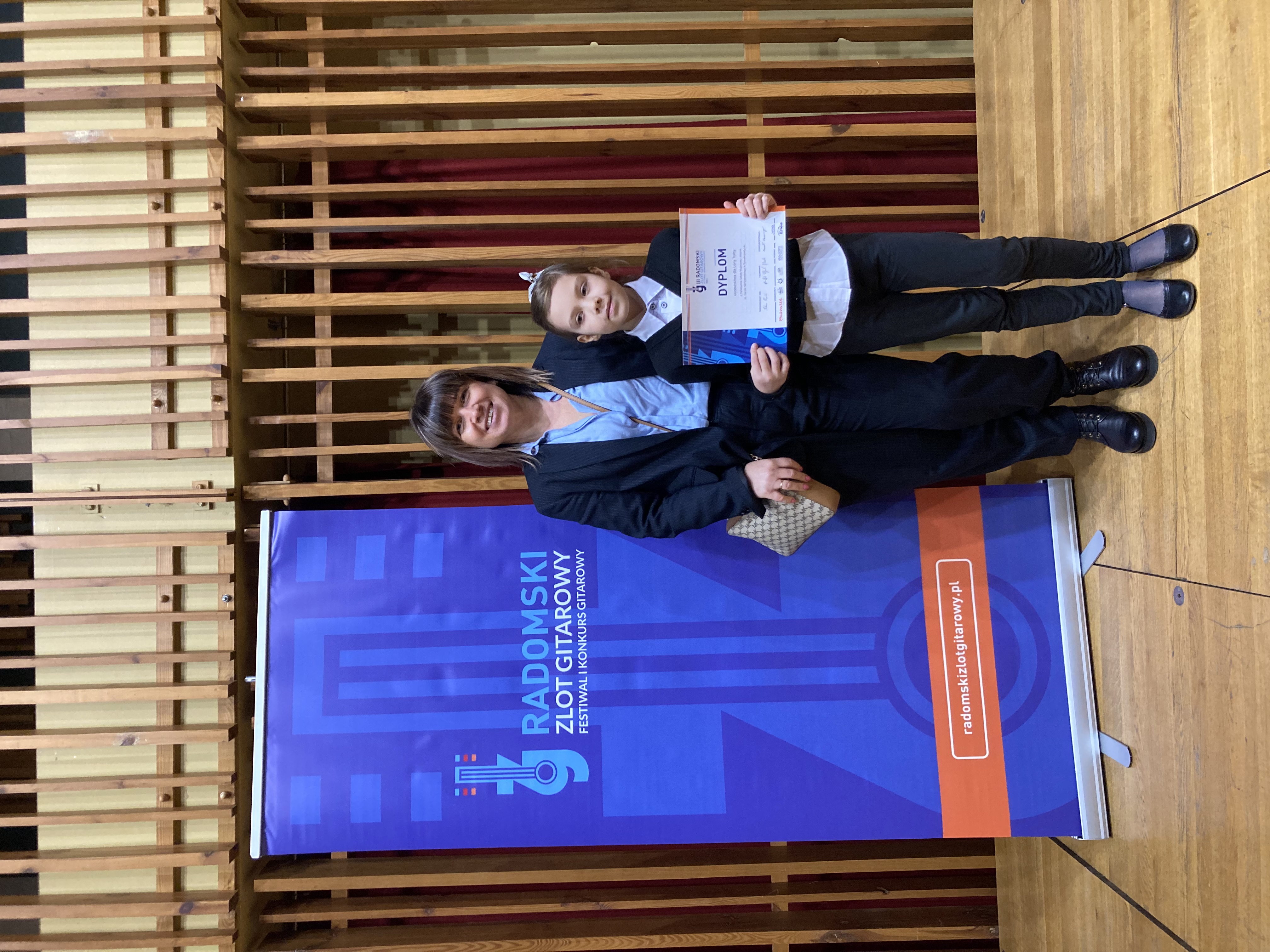 Zdjęcie przedstawia uczennicę Lenę Torba trzymającą dyplom, obok stoi nauczyciel gitary pani Iwona Grabowska. Na zdjęciu widać scenę na której stoi baner Radomskiego zlotu Gitarowego.