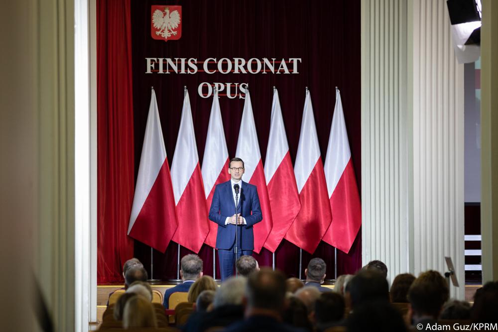 Premier Mateusz Morawiecki przemawia na scenie na tle flag Polski.