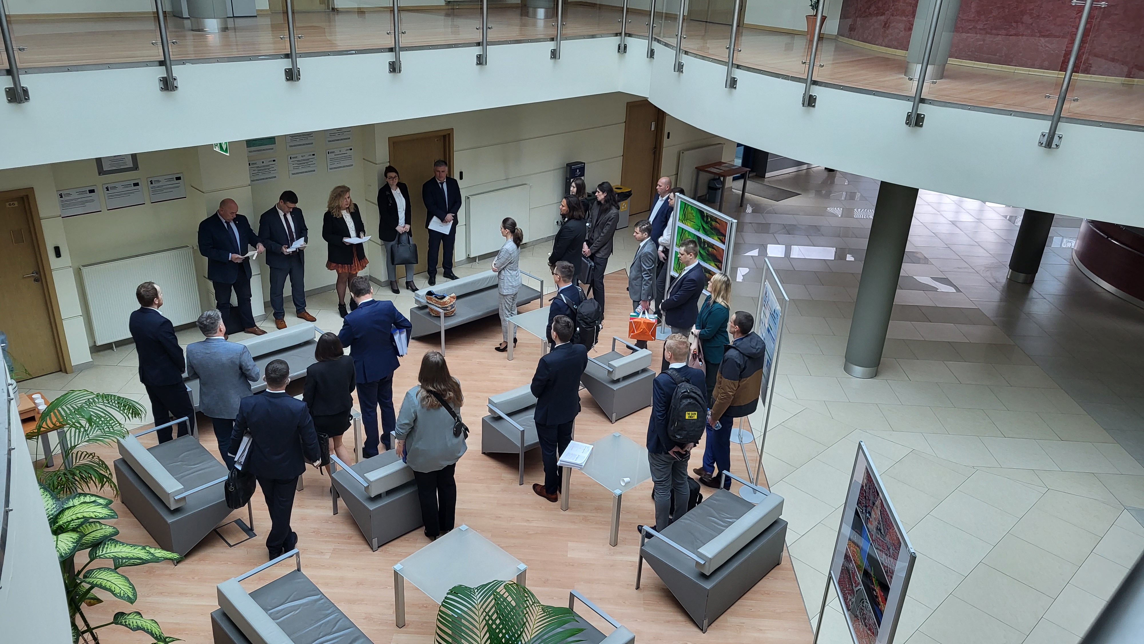 Zdjęcie przedstawia hol w siedzibie GUGiK przy ul. Olbrachta 94B z osobami przystępującymi do egzaminu słuchającymi wyjaśnień członków Komisji Egzaminacyjnej.