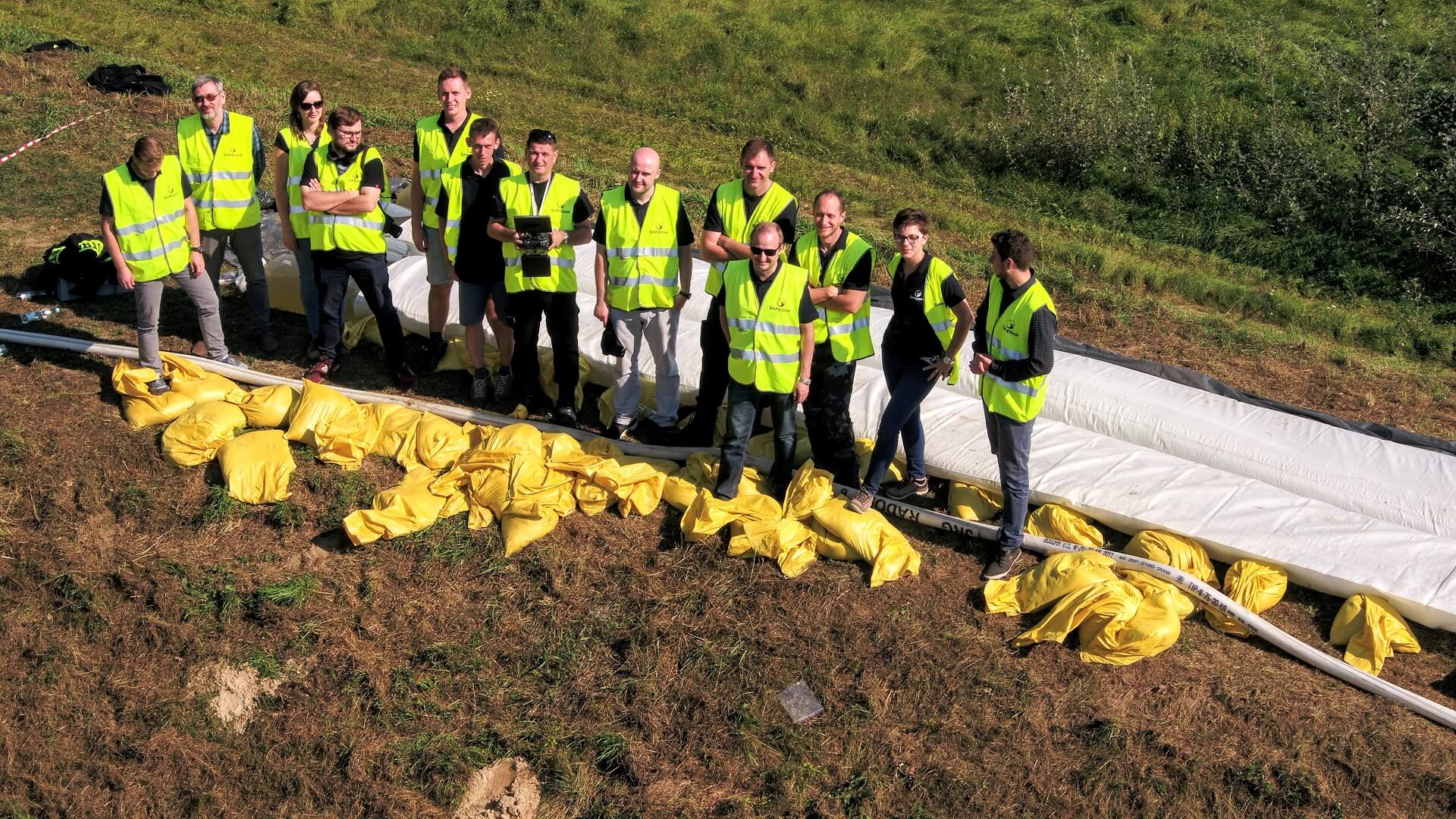 A group of thirteen from the SAFEDAM team, wearing yellow reflective vests, during maneuvers of the State Fire Service in Kozienice. On the ground a fire hose, yellow bags