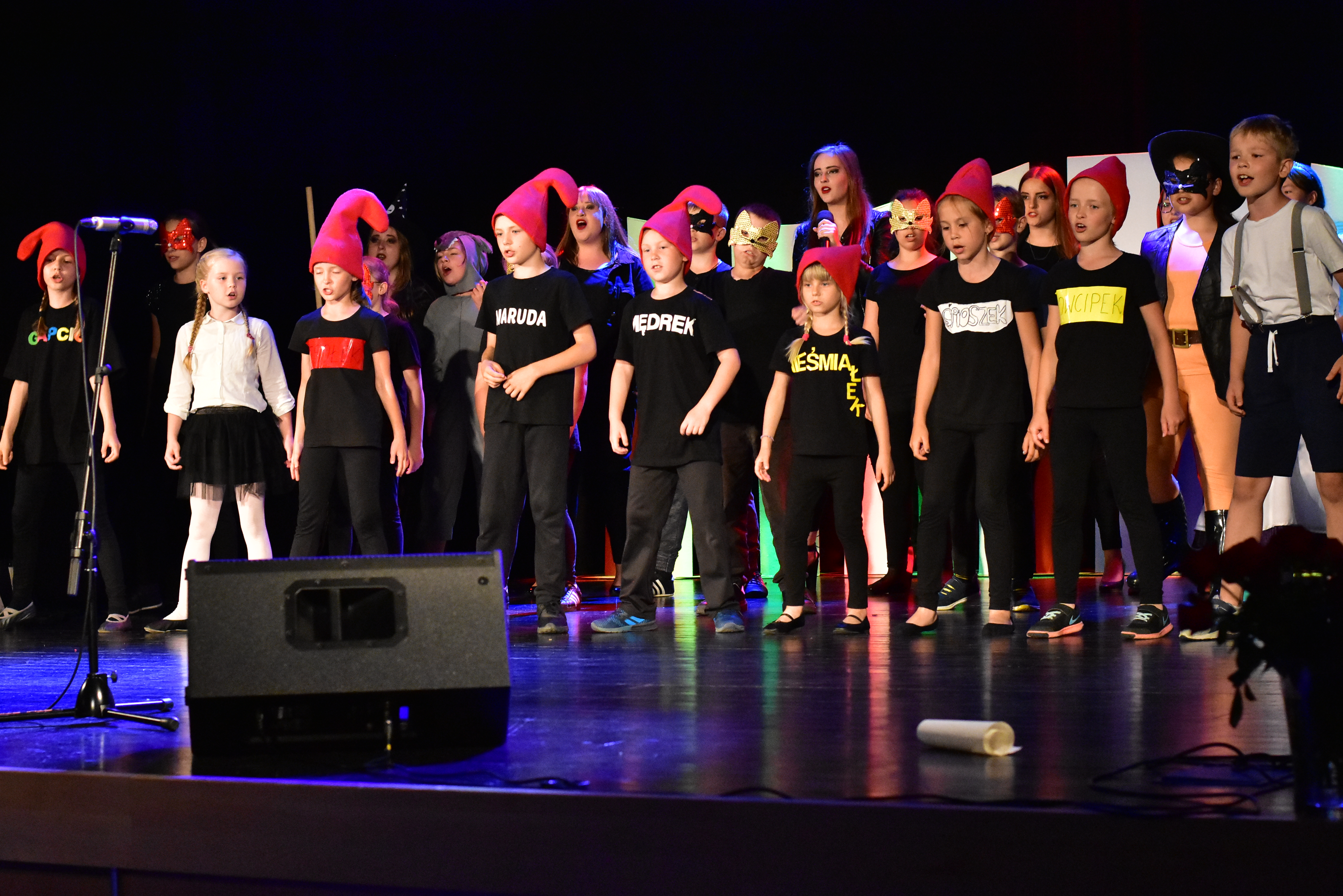 Zdjęcie przedstawia stojącą grupę dzieci. Większość z nich ma na sobie czerwone czapki oraz czarne stroje z napisami "Dowcipek" "Nieśmiałek" "Maruda".