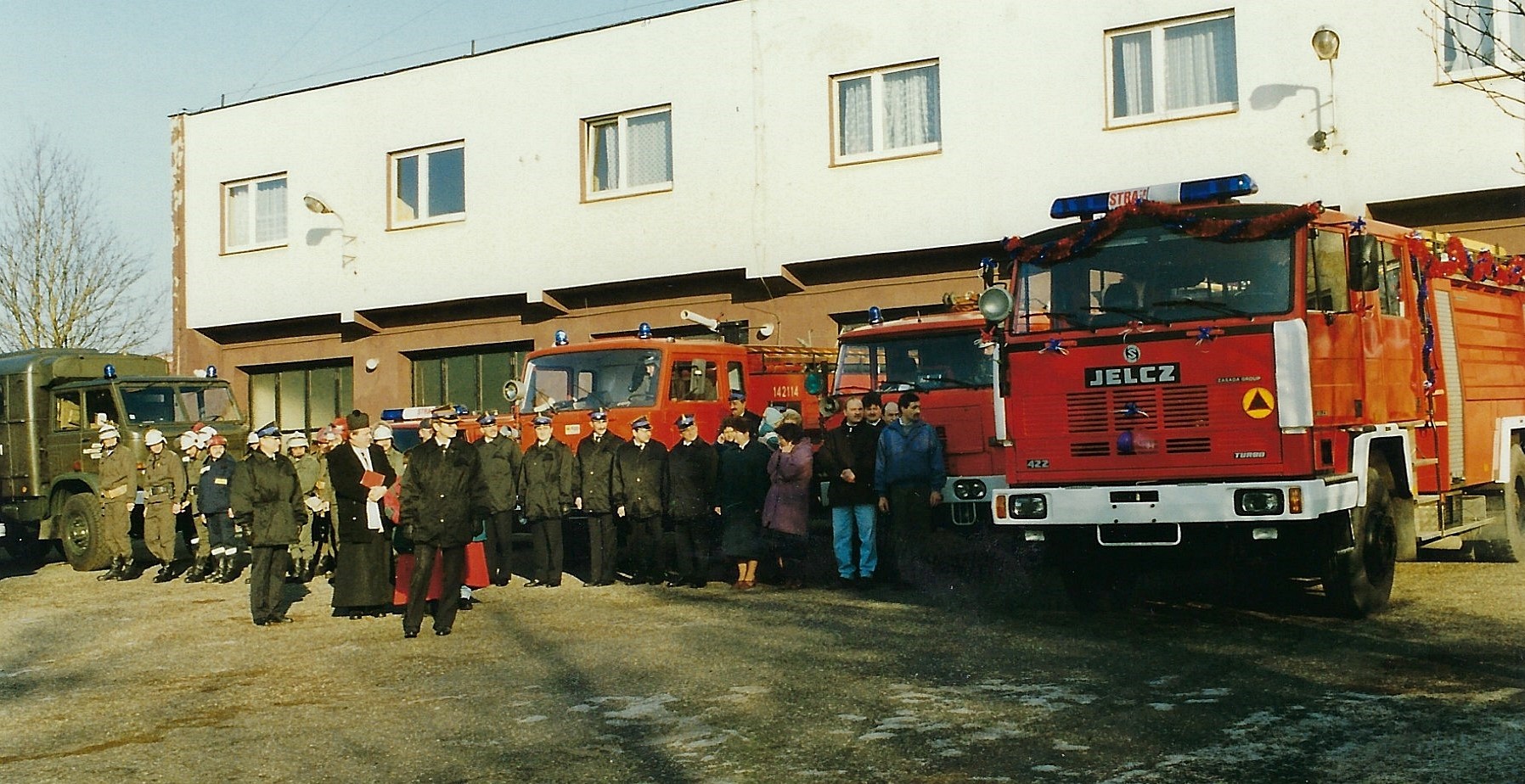 Na historycznym zdjęciu zbiórka strażaków oraz księdza podczas pierwszych odwiedzin kolędowych oraz poświęcenia nowego samochodu. W tle samochody pożarnicze oraz budynek Komendy.