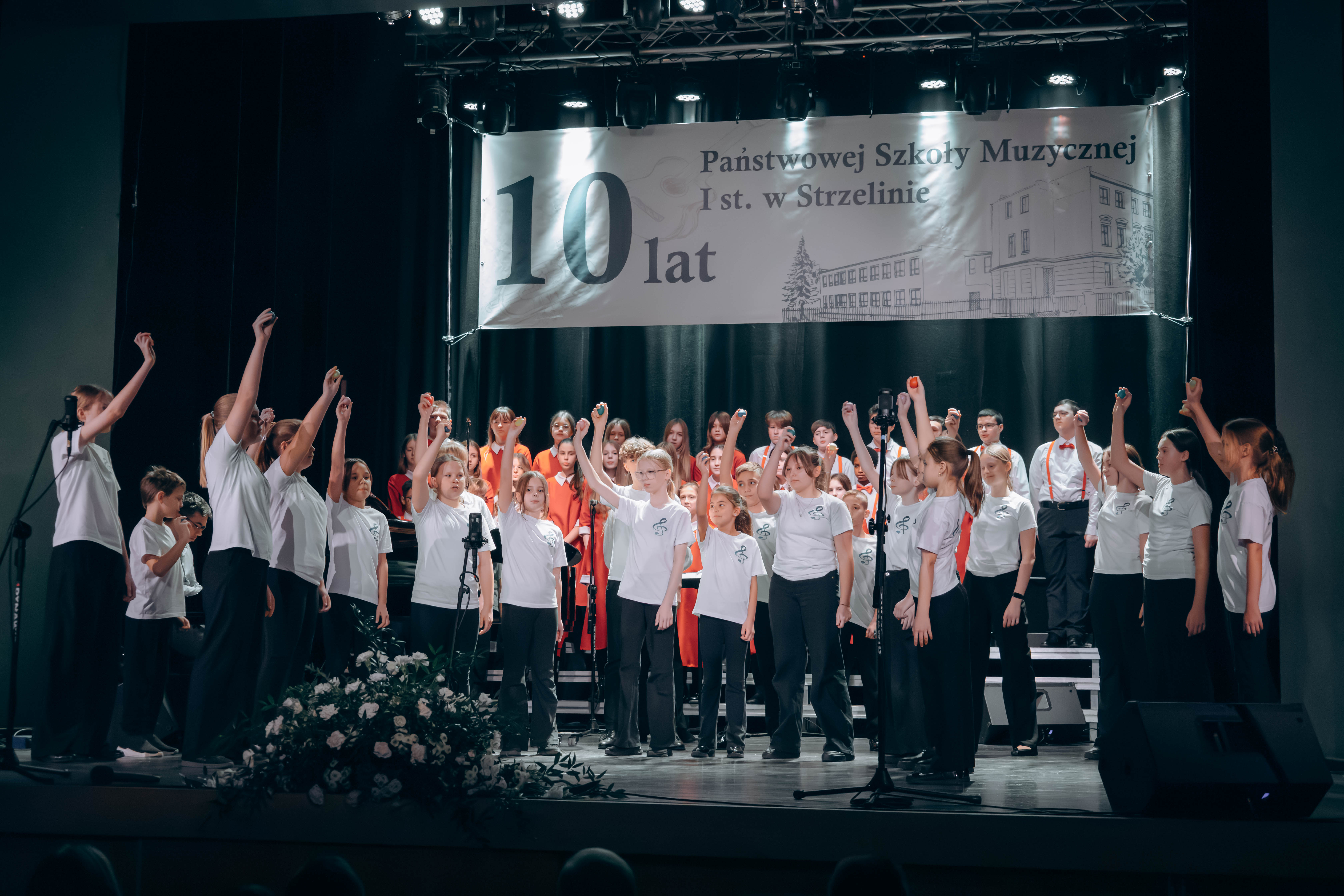 Na zdjęciu widoczna jest duża grupa tańczących na scenie dzieci. Za nimi stoi chór szkolny.