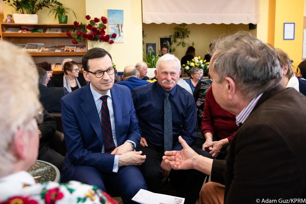 Premier Mateusz Morawiecki siedzi i rozmawia z seniorami.