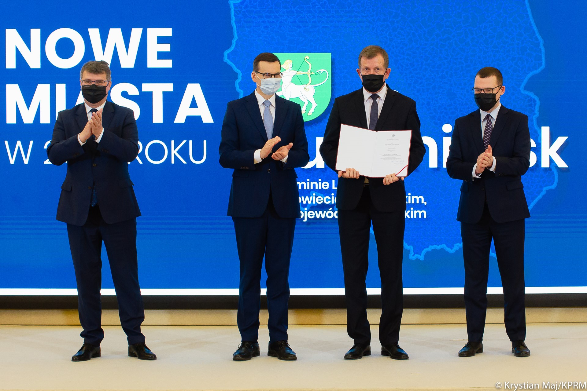 Premier wręcza akt nadania praw miejskich, obok stoi minister Paweł Szefernaker i minister Maciej Wąsik.