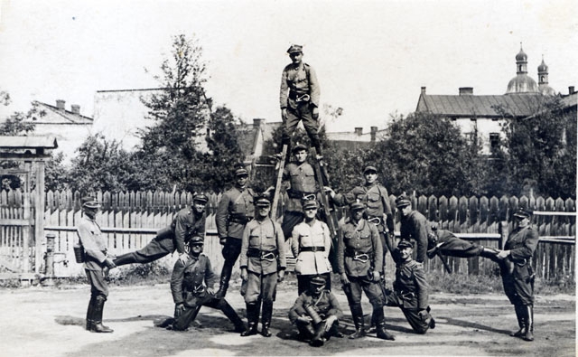 Zmiana służbowa Miejskiej Straży Pożarnej w Nowym Sączu w 1927 roku.stoi na placu. Dwóch wykonuje akrobacje jeden z nich stoi na rozłożonej drabinie. W tle widoczne budynki Nowego Sącza z wieżami Bazyliki Św. Małgorzaty.