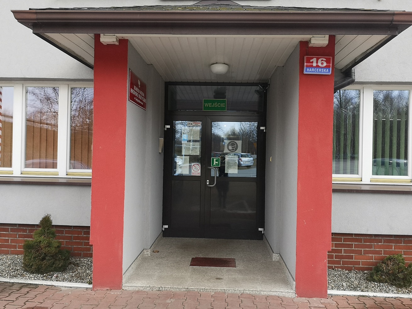 Zdjęcie przedstawia wejście główne do budynku. Na wprost znajdują się ciemne dwuskrzydłowe drzwi prowadzące do komendy. Drzwi mają przeszklenia, na przeszkleniach znajdują się informacje dla petentów, symbol otwierania drzwi na zewnątrz oraz symbol o zakazie palenia papierosów na terenie obiektu. Przed drzwiami znajduje się wycieraczka do butów. Ściany wokół wejścia są szare z pionowymi pasami w kolorze blado-czerwonym. Po obu stronach wejścia widać okna budynku komendy. Okna zasłonięte są pionowymi roletami. Lewe okno ma rolety brązowe a prawe zielone. Przed wejściem znajduje się chodnik wykonany z kostki brukowej w kolorze blado-czerwonym. 