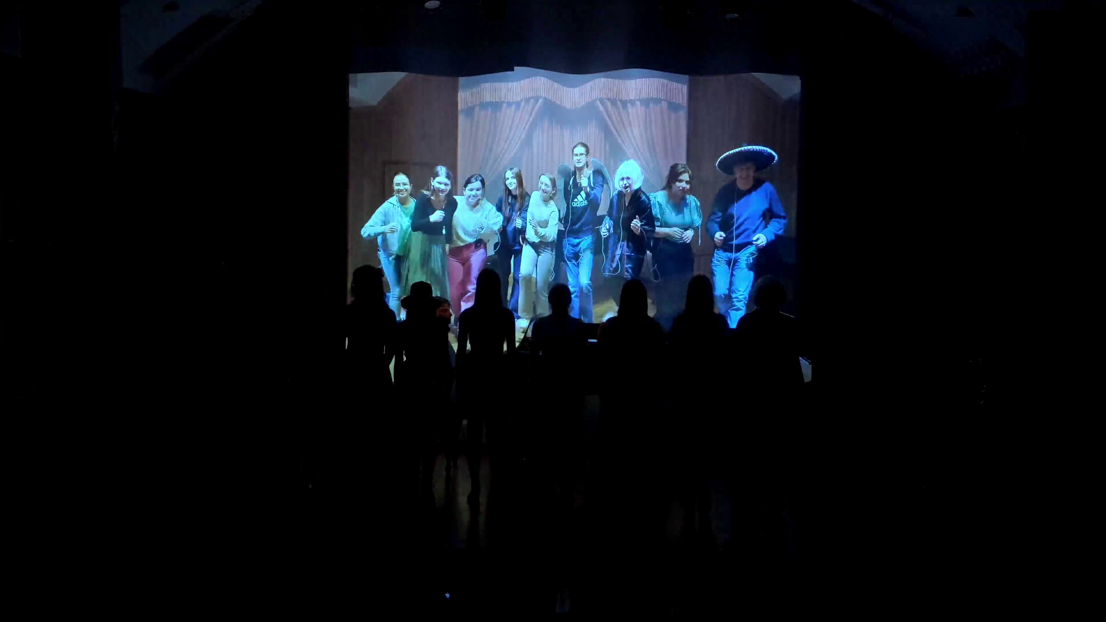 Zdjęcie. Na zaciemnionej estradzie stoją wykonawcy koncertu. Na wyświetlonym z tyłu zdjęciu widać ich sylwetki. Na zdjęciu utrwalony jest występ podczas wcześniejszej próby.