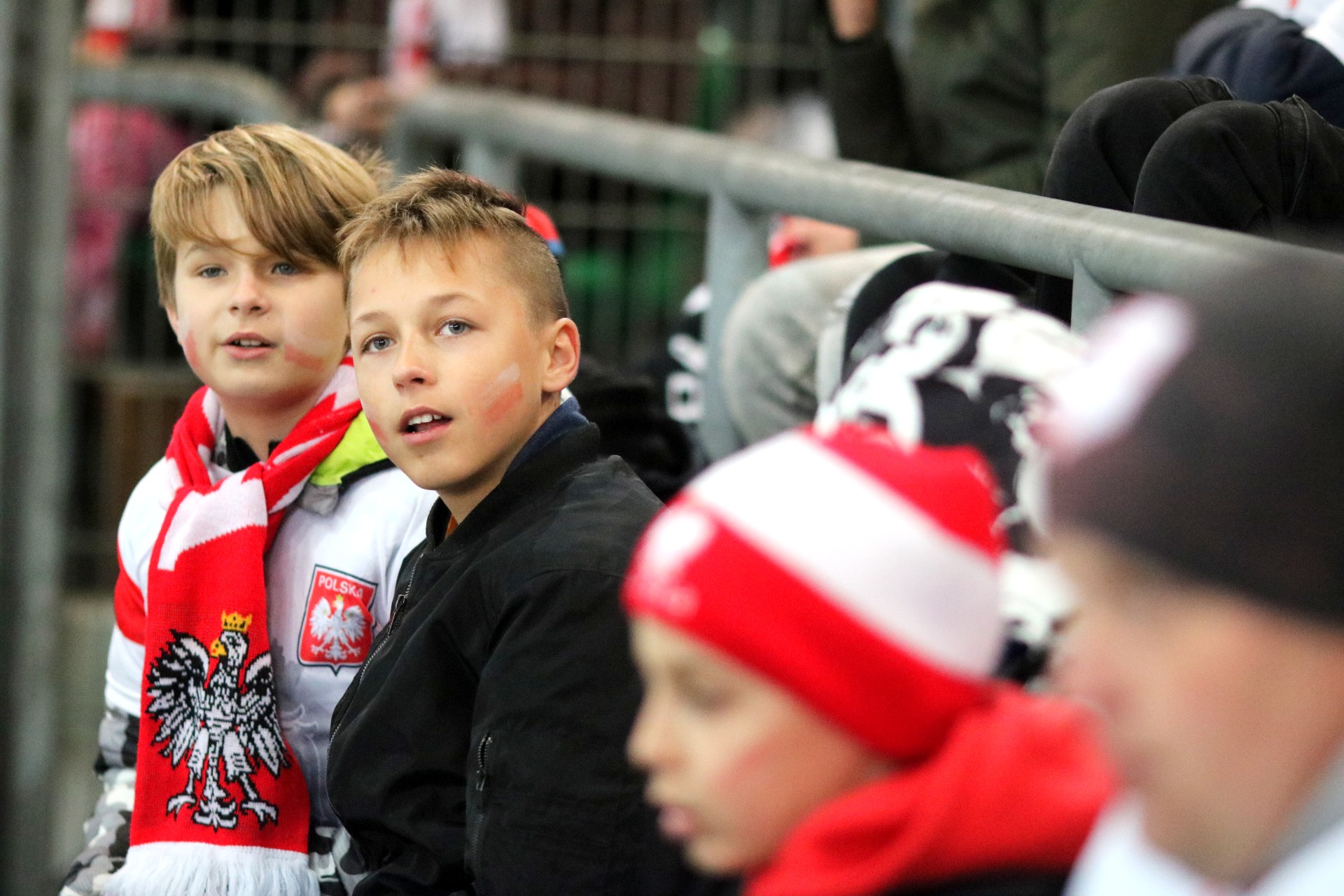Grupa chłopców siedzi na stadionie, kilku ma biało-czerwone szaliki i czapki.